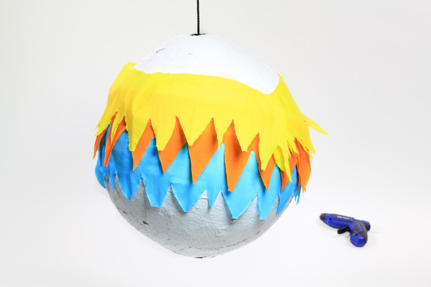 Auf dem zweiten Bild ist die Piñata-Kugel mit dem Krepppapier verziert. Daneben liegt eine Heißklebepistole.