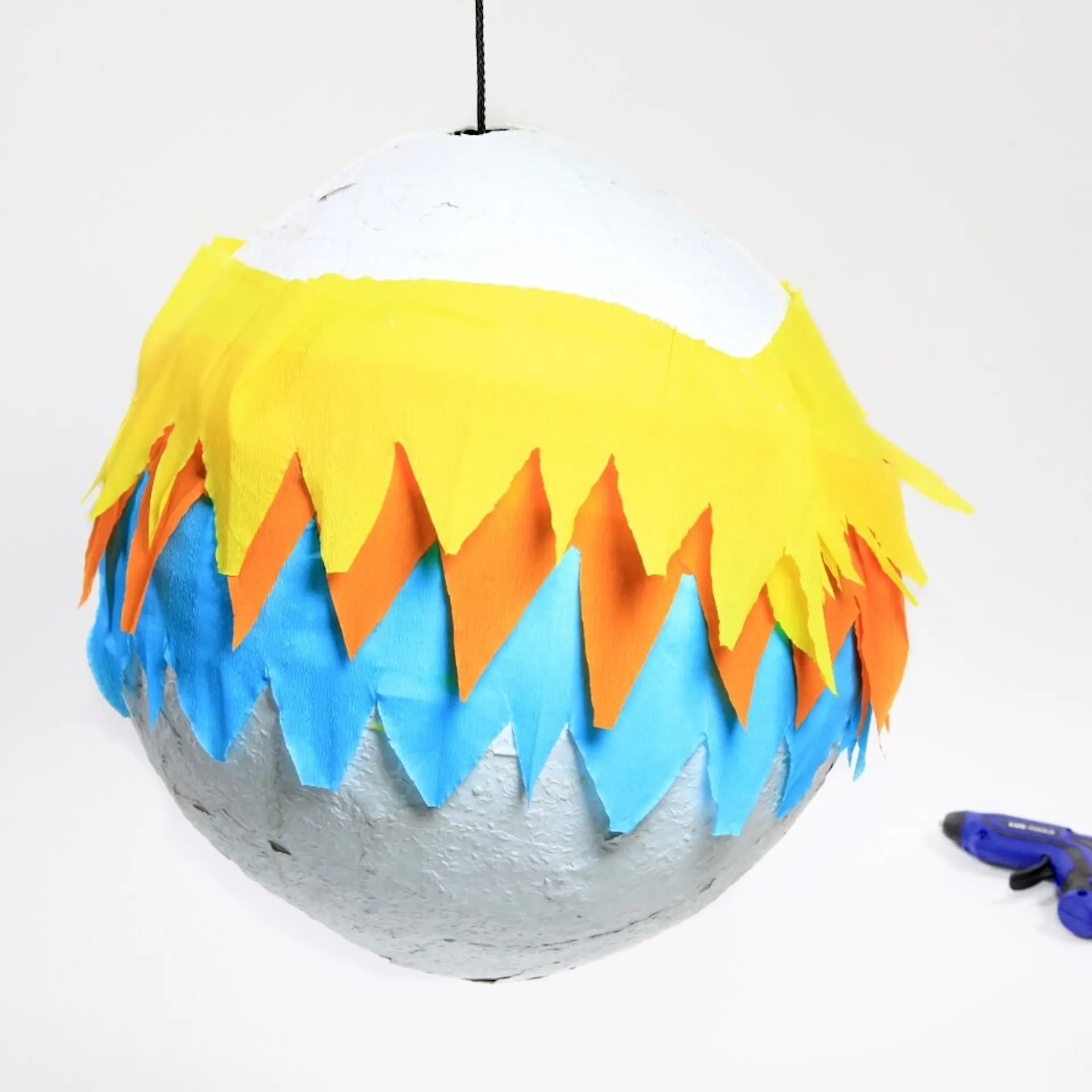 Auf dem zweiten Bild ist die Piñata-Kugel mit dem Krepppapier verziert. Daneben liegt eine Heißklebepistole.