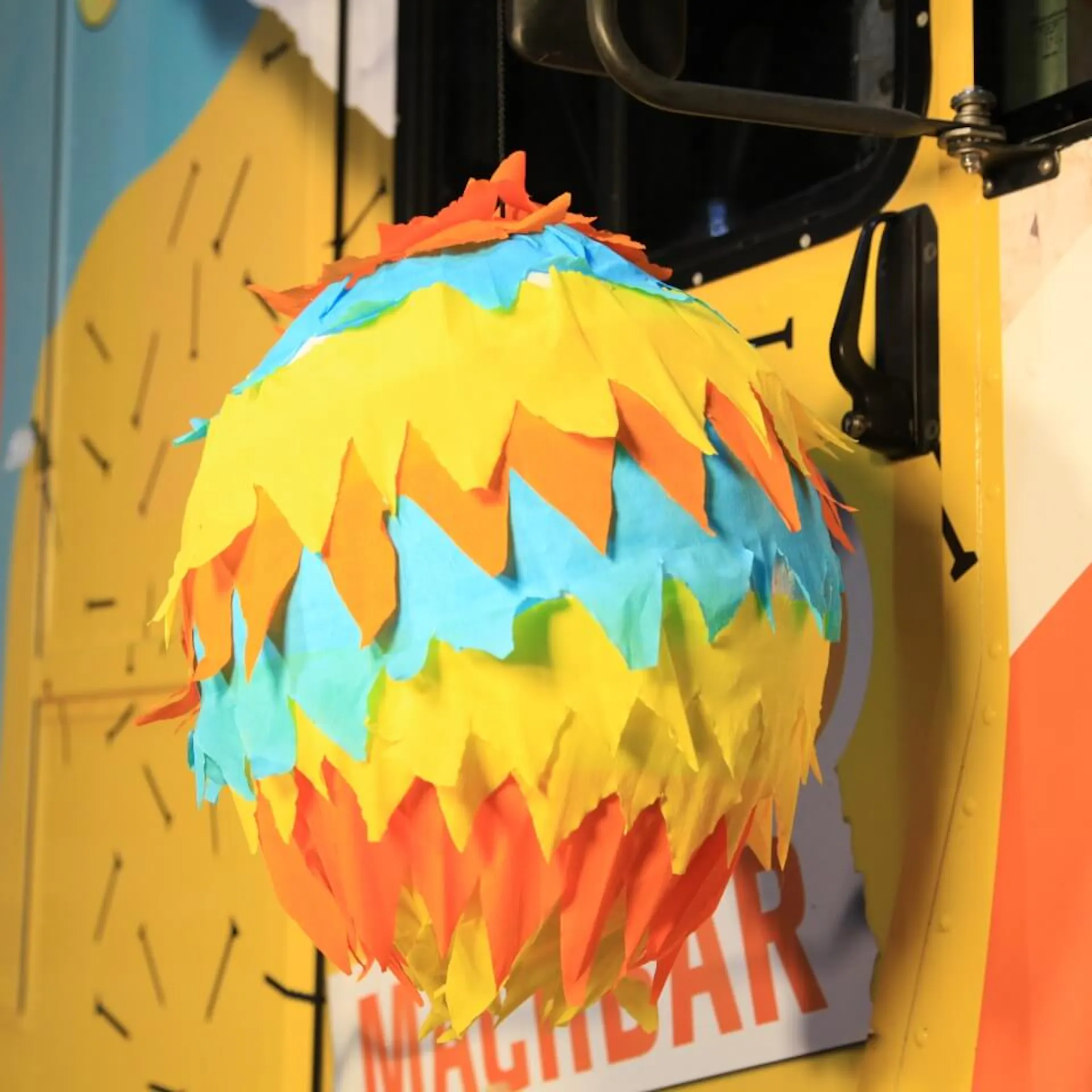 Die fertige Piñata ist vollständig mit buntem Krepppapier geschmückt. Sie hängt vor einer gelben Wand.