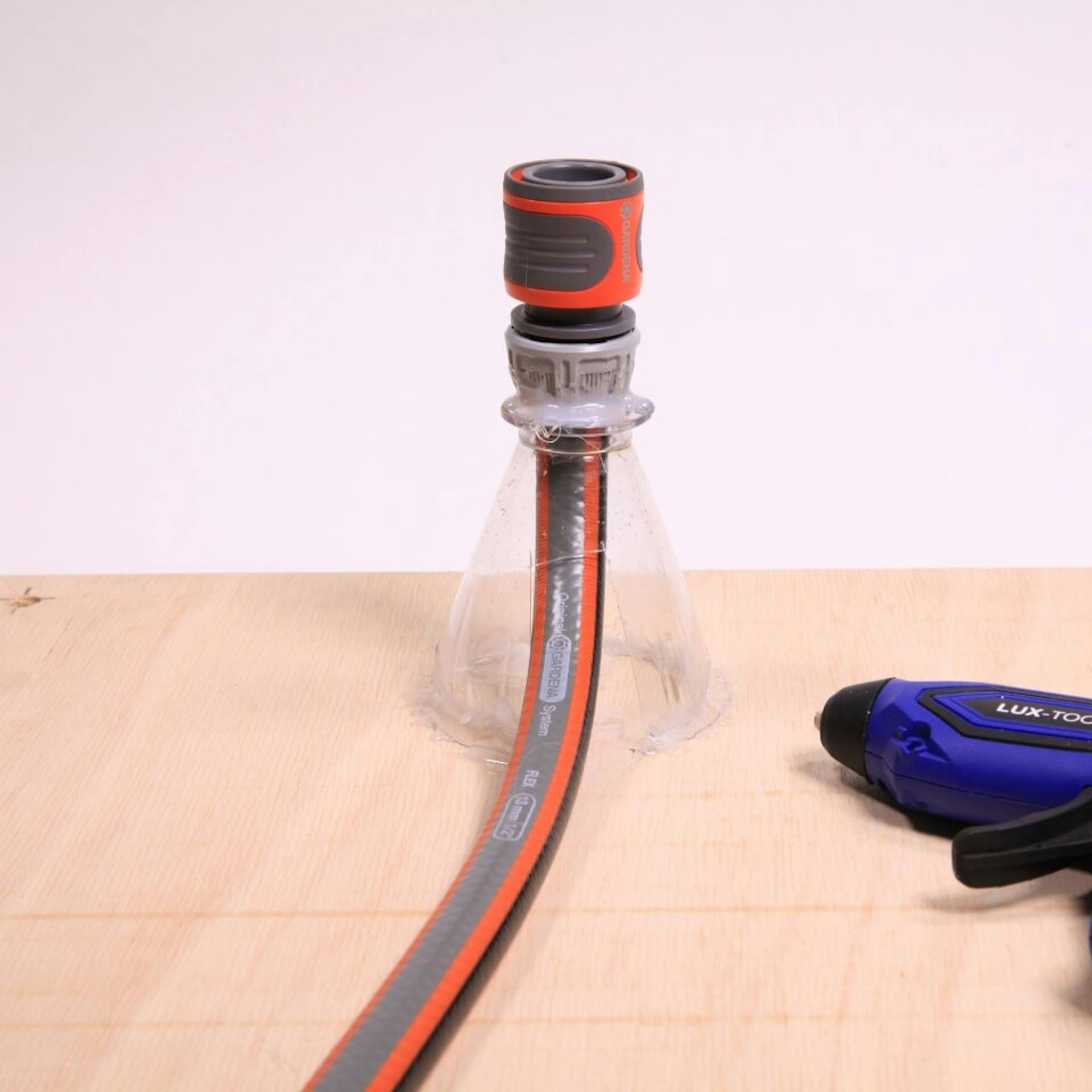 Ein Gartenschlauch, ein darauf aufgezogener Flaschenhals, festgeklebt auf einer Holzplatte. Daneben liegt eine Heißklebepistole.