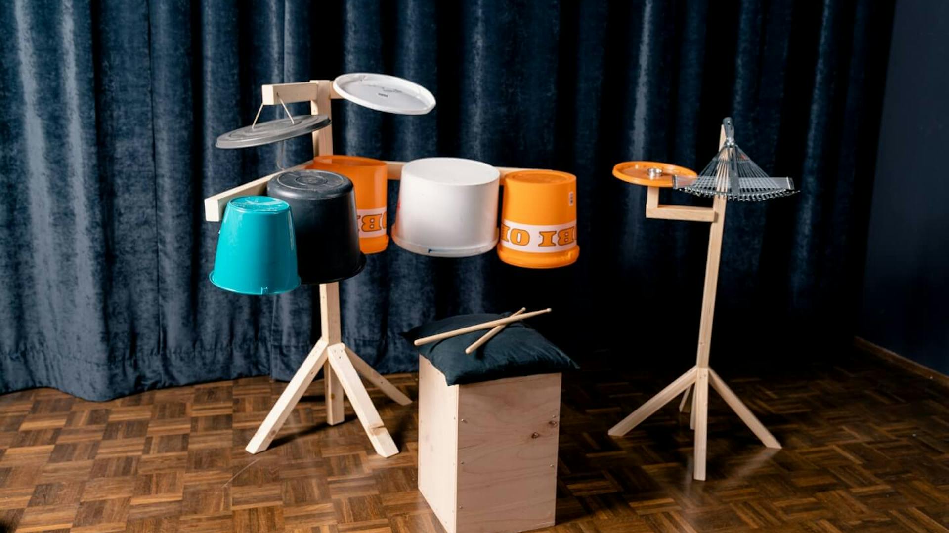 Auf einem Holzboden stehen zwei Ständer aus Holz sowie eine Holzkiste. An den Ständern sind mehrere Eimer in verschiedenen Farben befestigt. Auf der Holzkiste liegen Drumsticks sowie ein schwarzes Kissen. Im Hintergrund ist eine dunkle Samtgardine zu sehen.