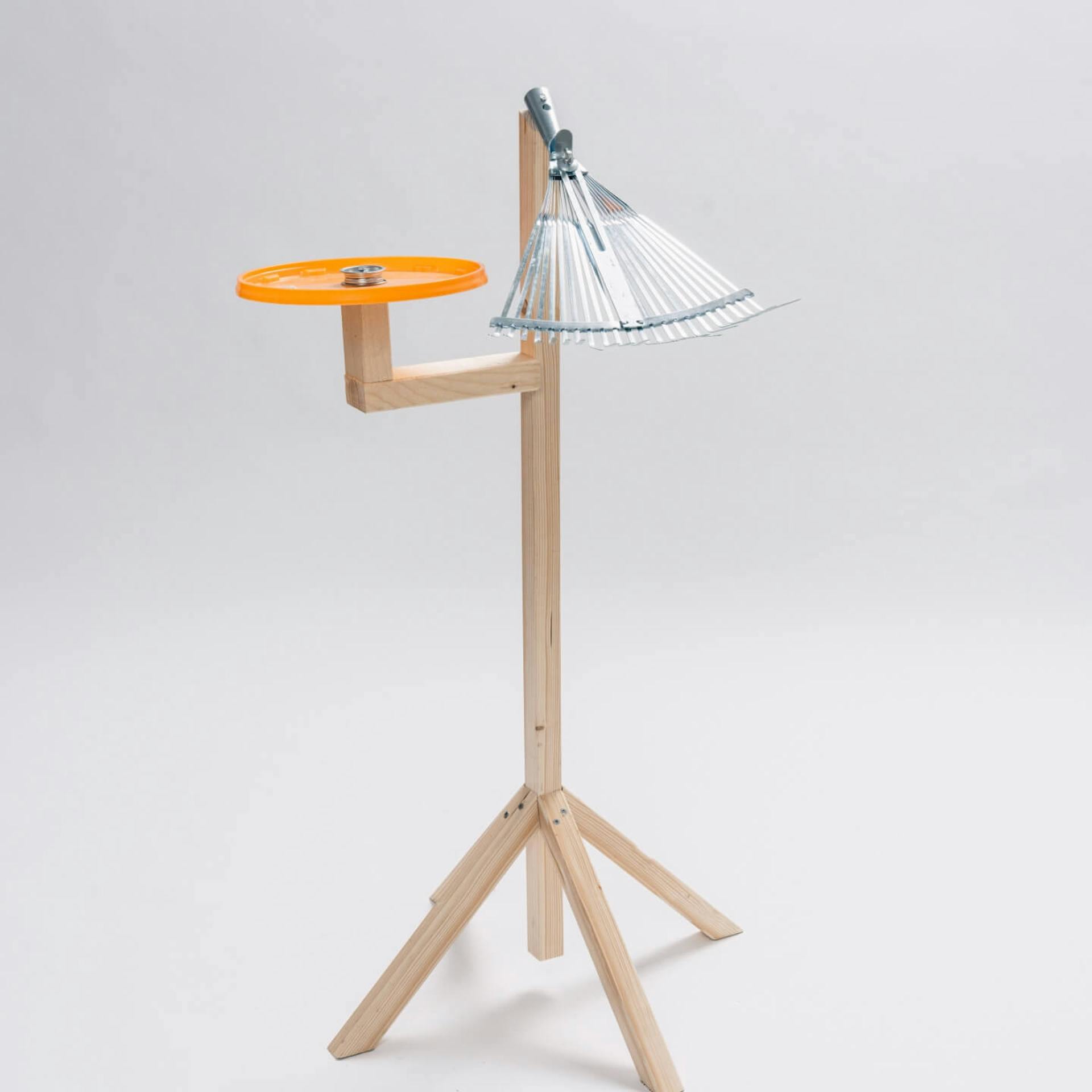 Es ist ein Ständer aus Holz zu sehen an welchem vier Holzfüße befestigt sind. Am oberen Teil des Ständers ist an einer Seite ein orangener Eimerdeckel befestigt, an der anderes Seite ein Rechen.