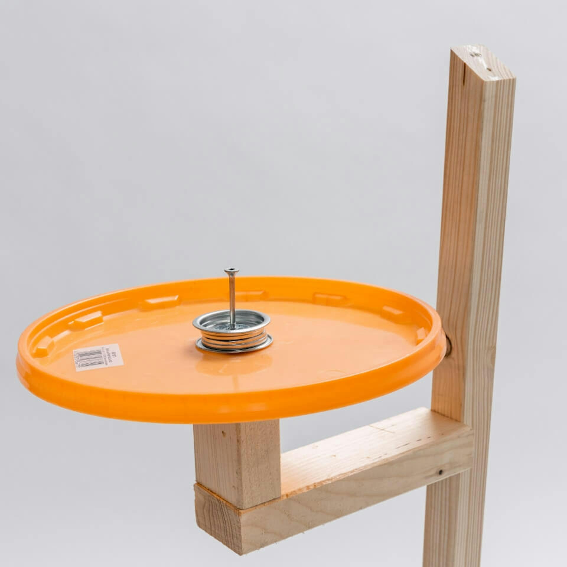 Es ist eine Halterung aus Holz zu sehen, an welcher ein orangener Eimerdeckel mit einer Schraube befestigt ist.