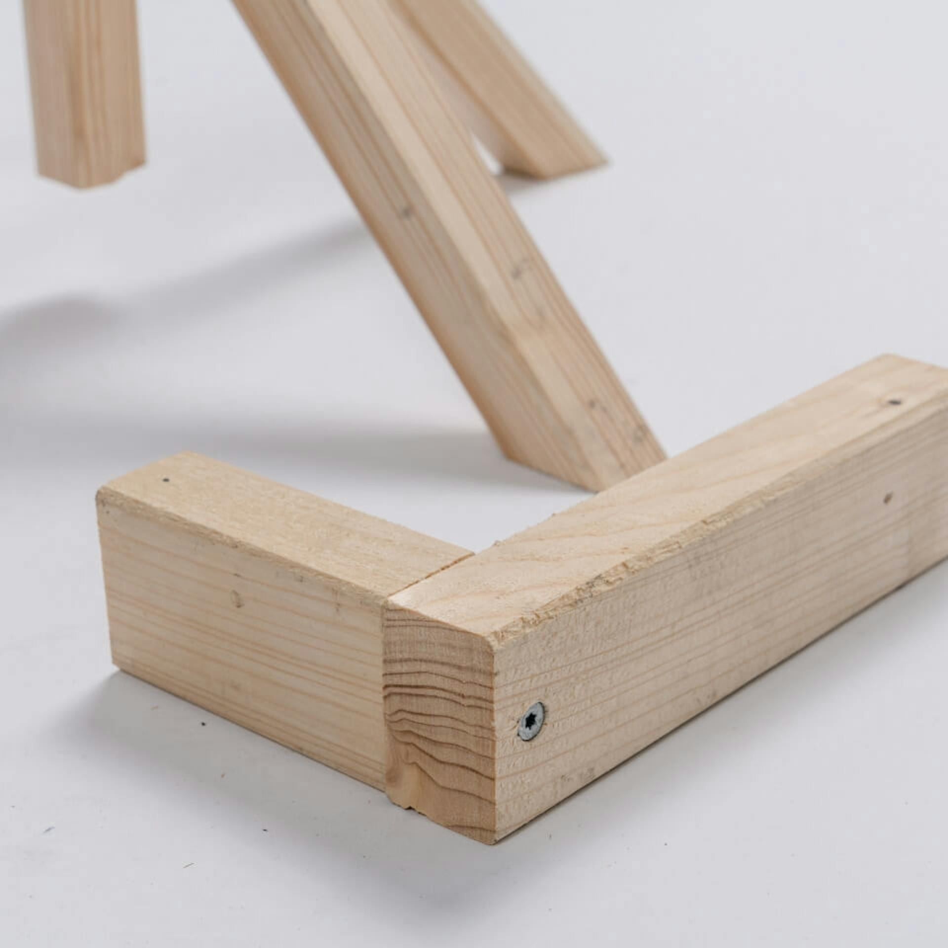 Im Hintergrund ist ein Holzständer zu sehen. Davor liegt auf einem hellen Untergrund ein Kantholz in L-Form.