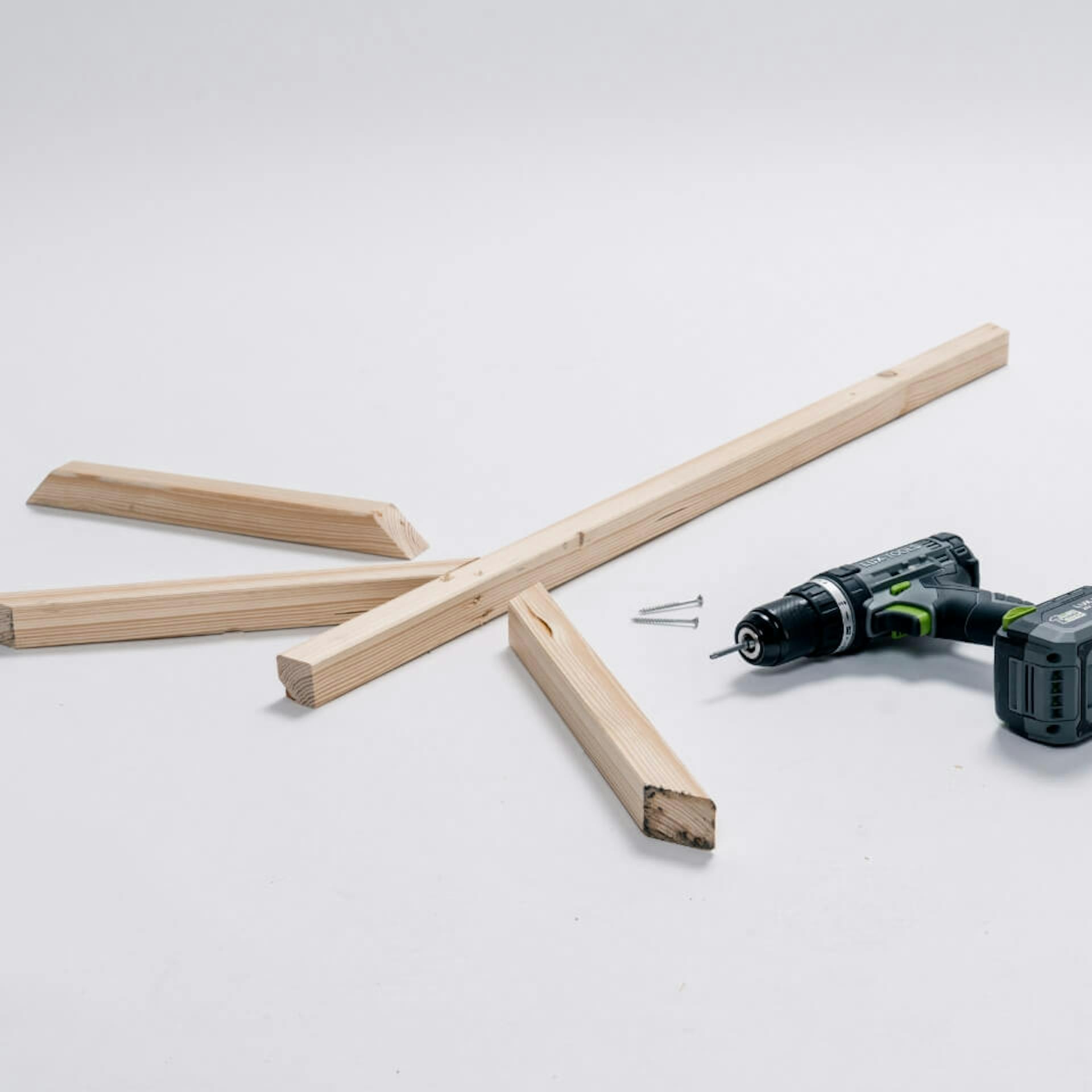 Auf einem hellen Untergrund liegt ein längeres Kantholz sowie drei kürzere Kanthölzer. Daneben liegen zwei Schrauben und ein Akkubohrer.