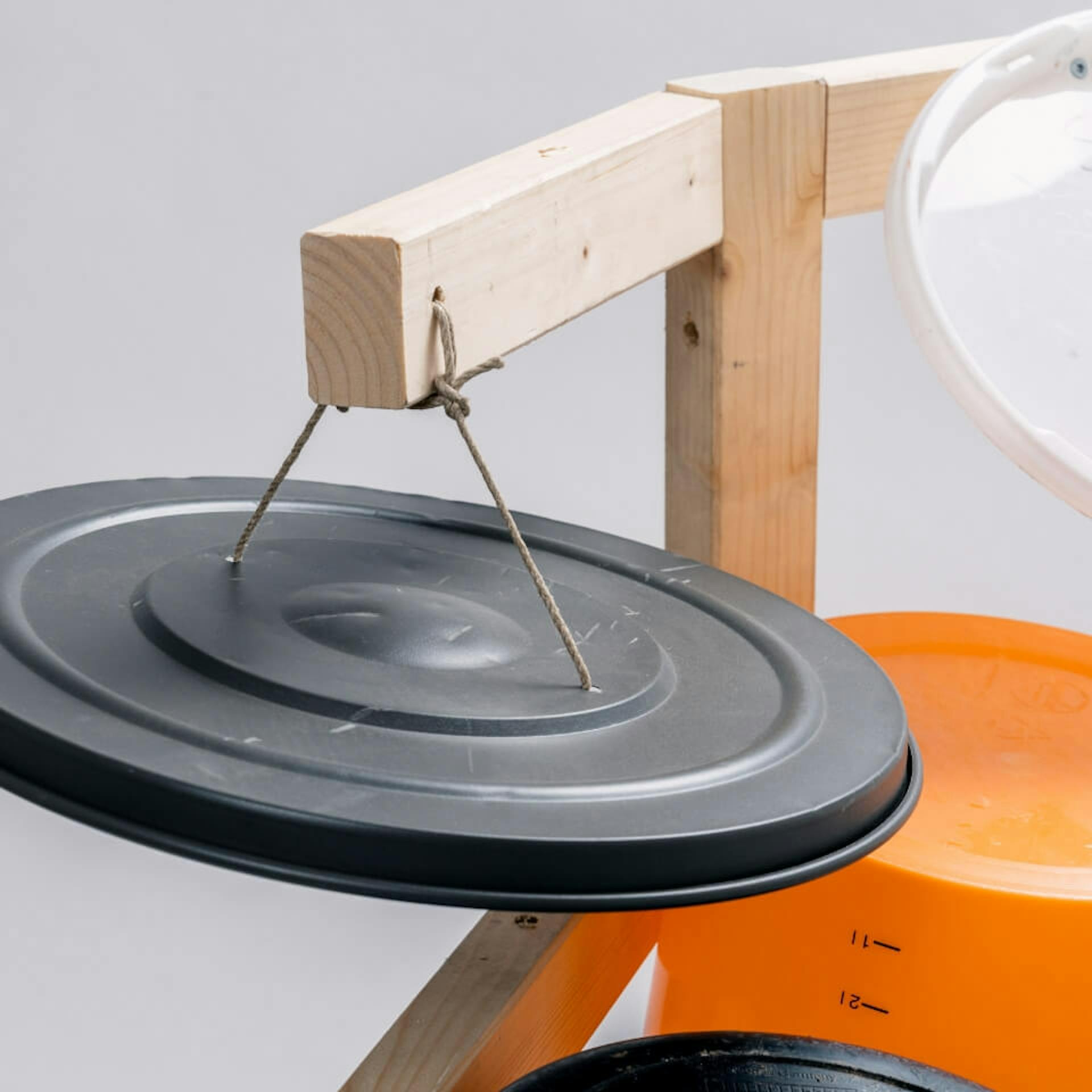 Es ist der obere Teil eines Ständers aus Holz zu sehen, an welchem zwei Kanthölzer befestigt sind. An den Kanthölzern sind jeweils Eimerdeckel befestigt.