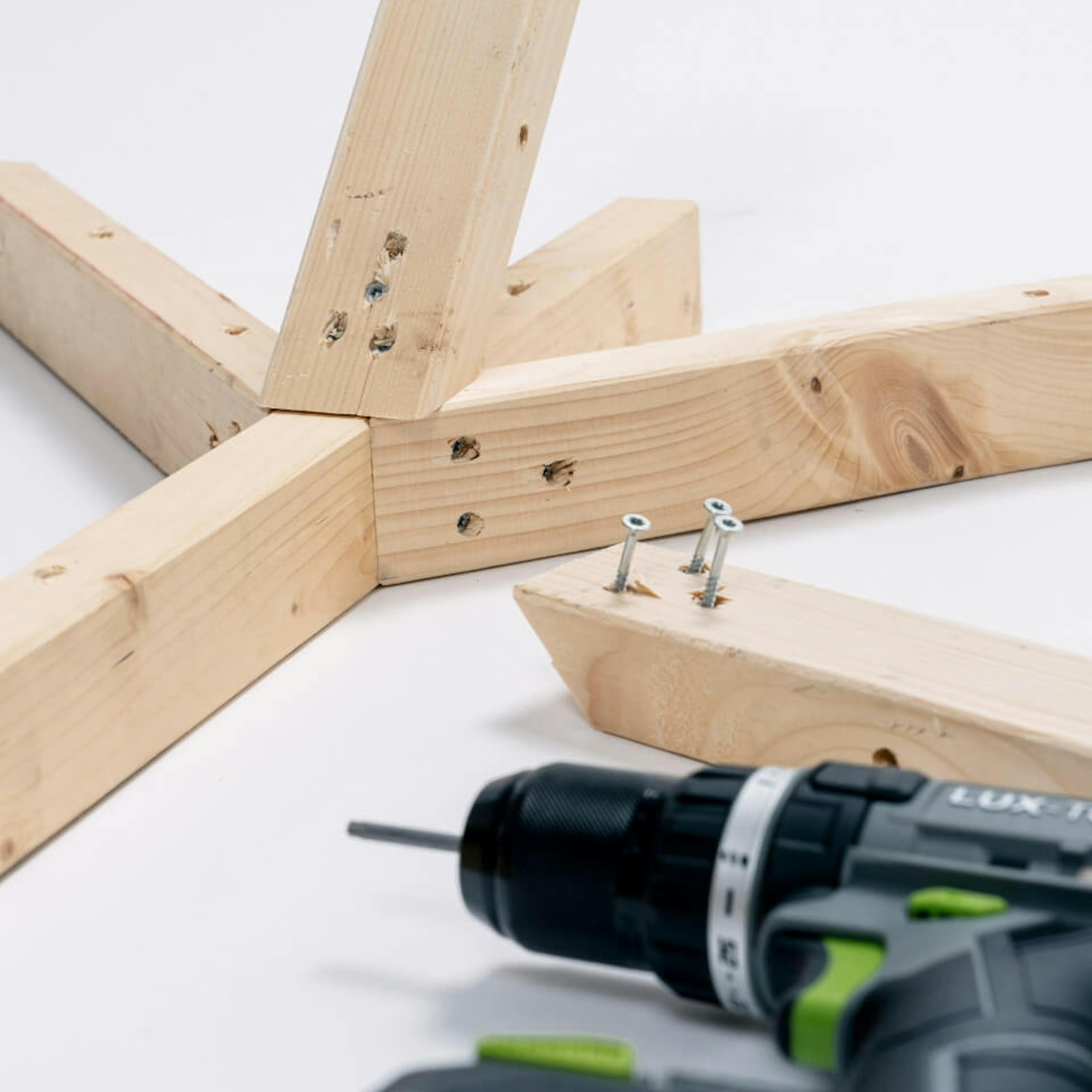 Auf einem hellen Untergrund liegt ein Kantholz, an welchem drei kürzere Kanthölzer befestigt sind. Davor liegt ein Kantholz, aus welchem drei Schrauben hervorragen. Im Vordergrund ist ein Akkubohrer zu sehen.