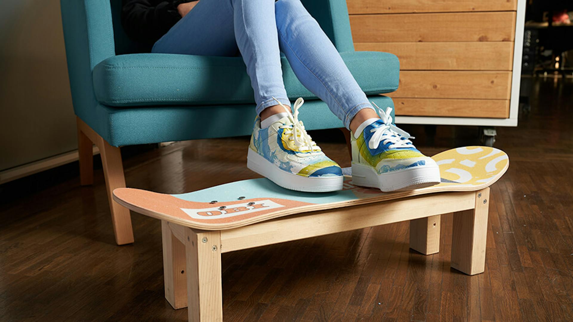 Eine jüngere Person sitzt in einem Sessel und hat die Beine auf den Skateborad Hocker gelegt.