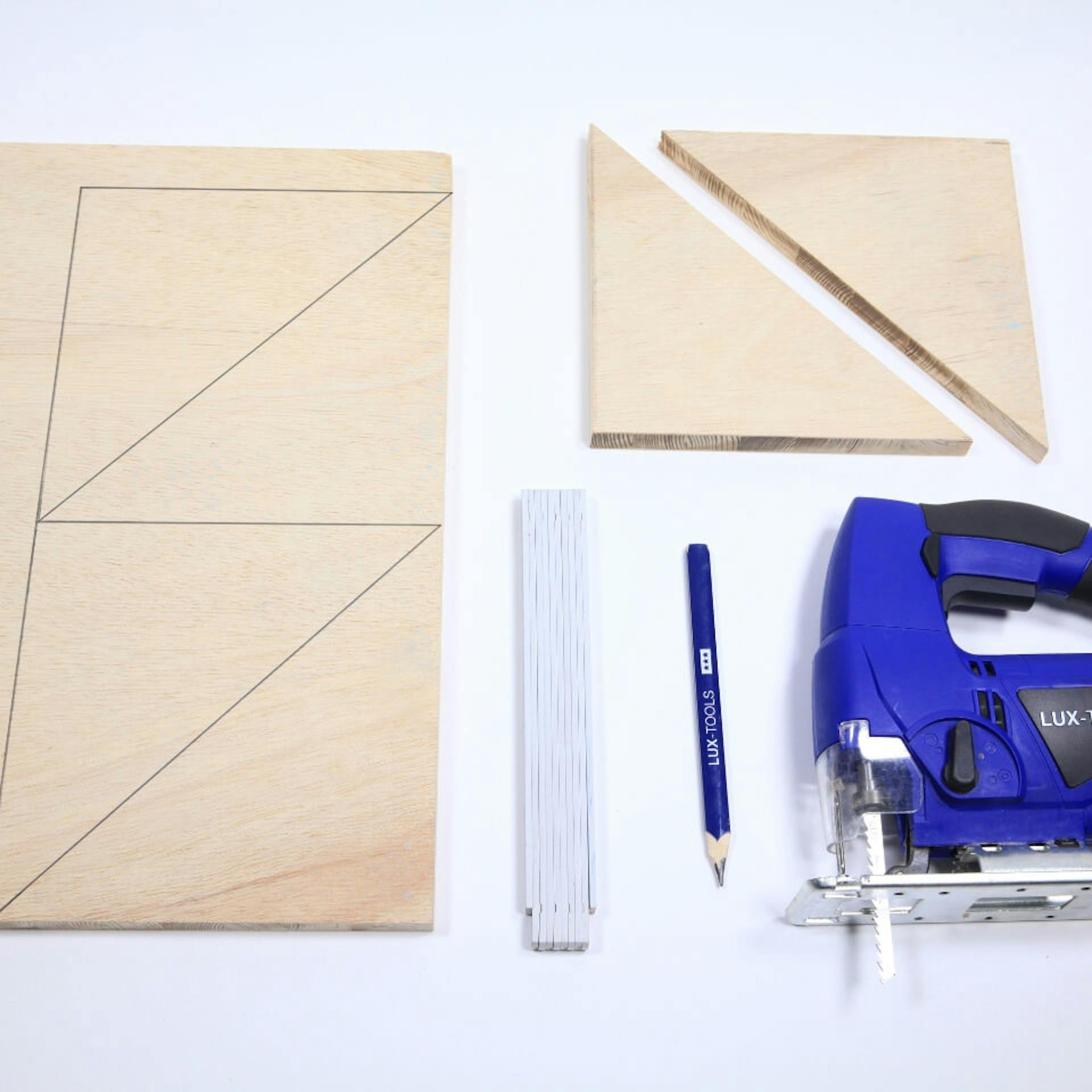 Es ist eine Holzplatte mit Bleistiftzeichnungen zu sehen. Daneben liegen zwei dreieckige Holzelemente, ein Zollstock sowie ein blauer Bleistift und eine Stichsäge.