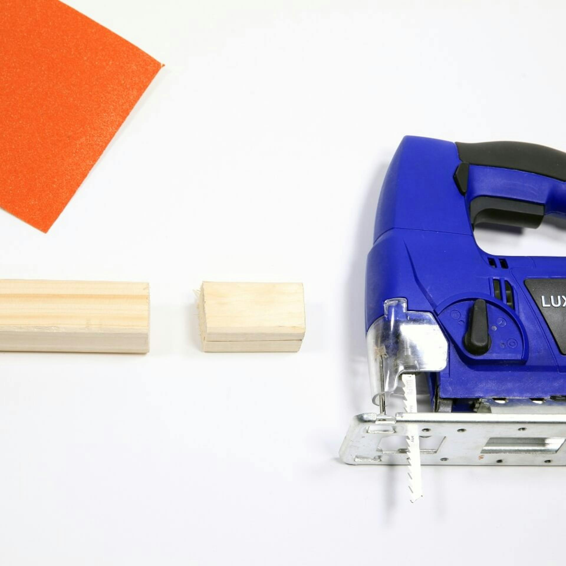 Es ist ein Kantholz zu sehen, daneben liegt ein kleines Holzstück. Neben dem Holz ist ein Stück Schleifpapier sowie eine blaue Stichsäge zu sehen.