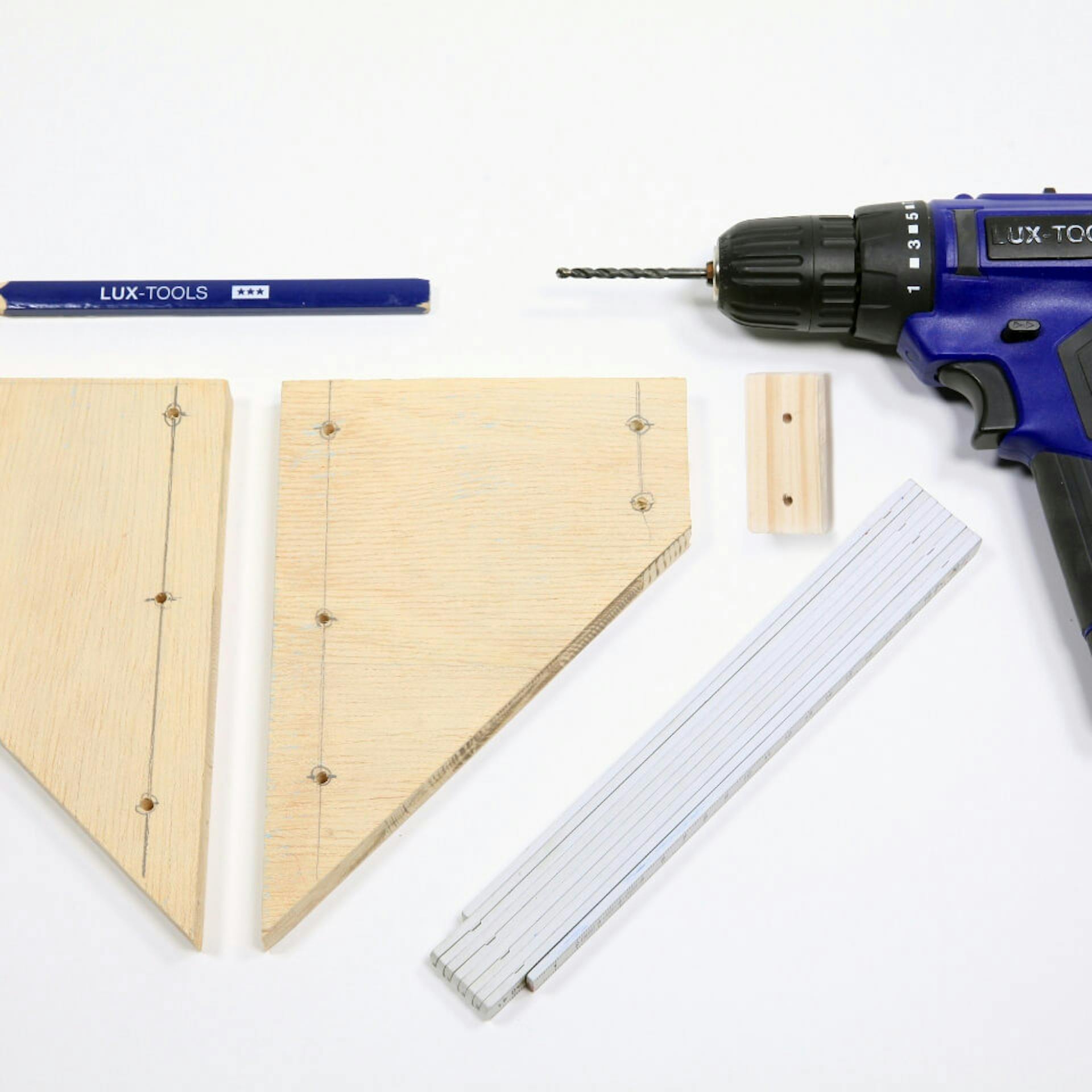 Es sind drei Holzelemente mit Bleistiftzeichnungen zu sehen. Daneben liegt ein Zollstock sowie ein blauer Bleistift und eine Stichsäge.