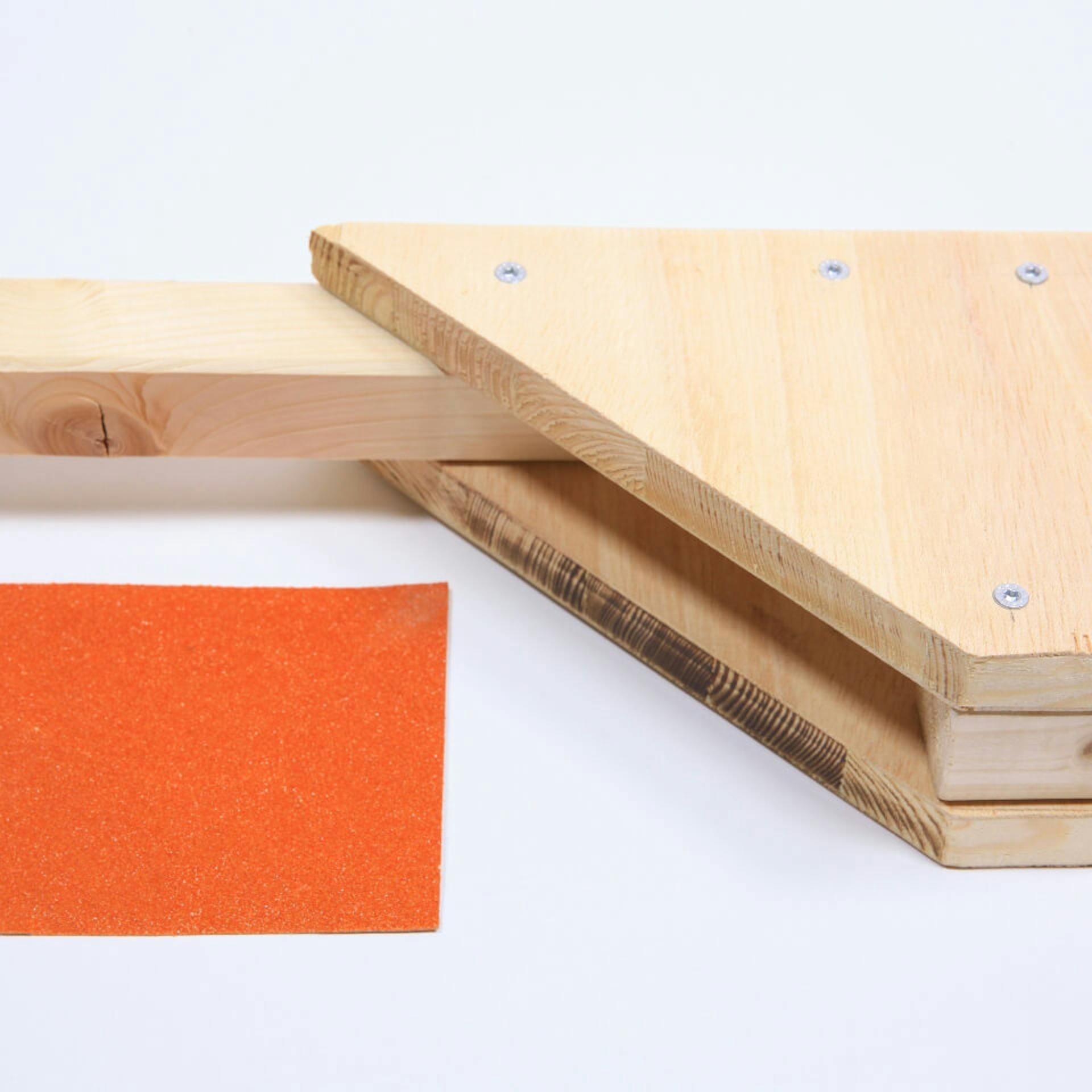 Es sind mehrere Holzelemente zu sehen, welche durch Schrauben miteinander verbunden sind. Daneben liegt ein Stück rotes Schleifpapier.
