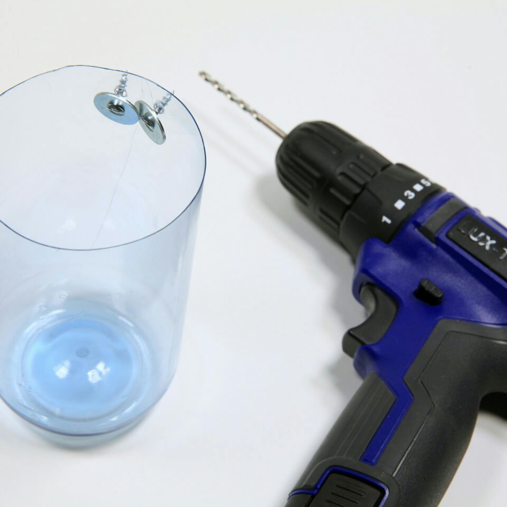 Es ist die untere Hälfte einer Plastikflasche zu sehen. Am Rand der Flasche sind zwei Schrauben mit Unterlegscheiben befestigt. Daneben liegt ein blauer Akkuschrauber.