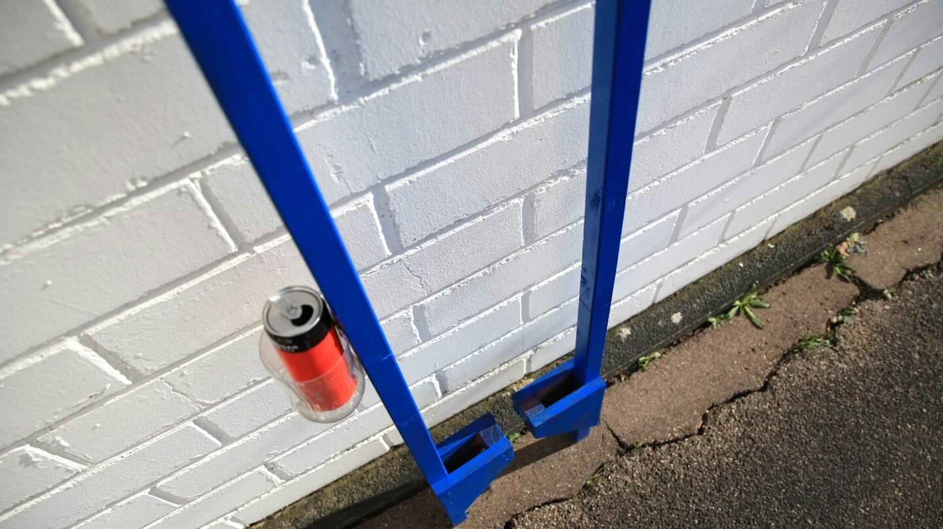 Es sind zwei blaue Holzstelzen zu sehen, welche an einer weißen Wand angelehnt sind. An einer Stelze ist ein Getränkehalter befestigt, in welchem sich eine Dose befindet.