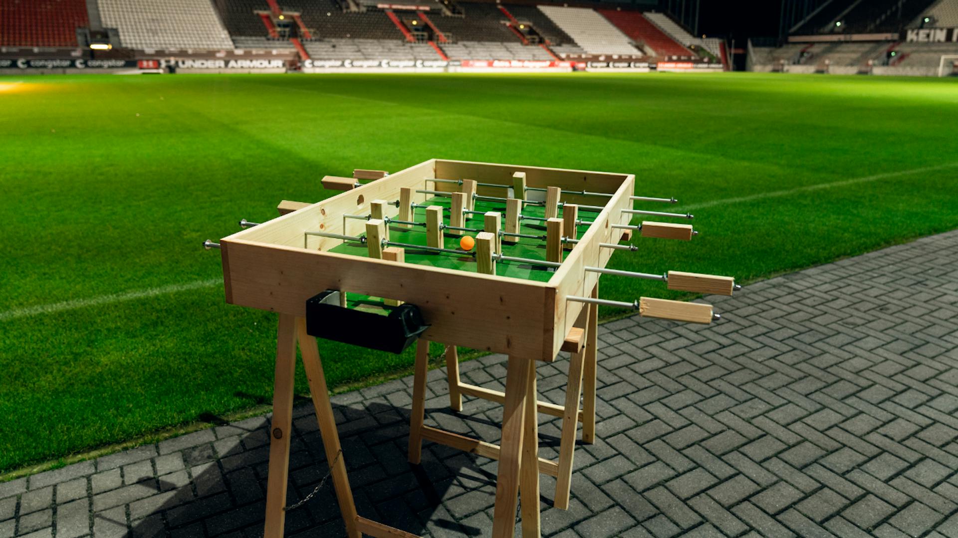 Tischkicker steht am Rasenrand eines großen Fußballstadions.