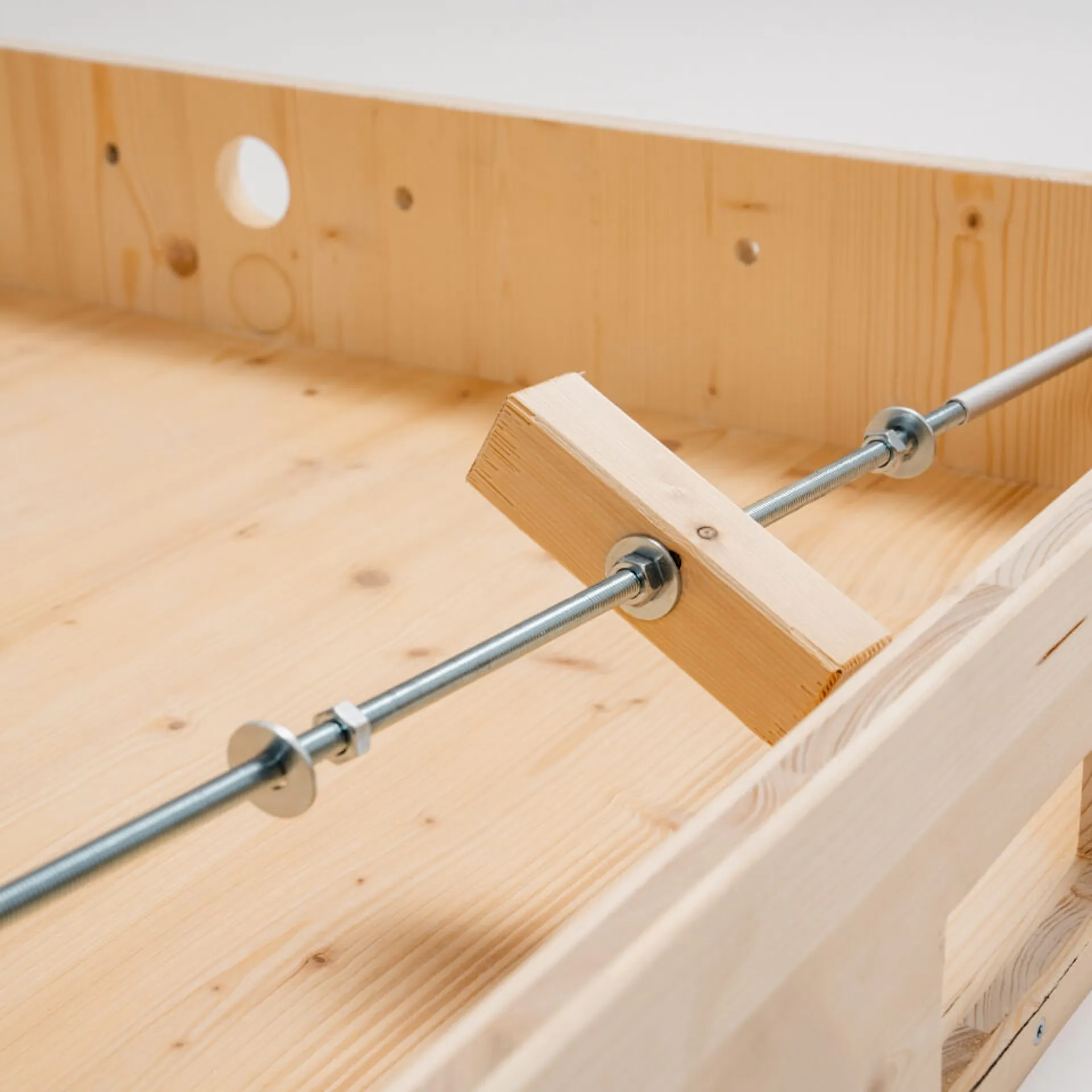 Závitová tyč s hranolem, upevněná maticemi a podložkami, vložena do dřevěné konstrukce.