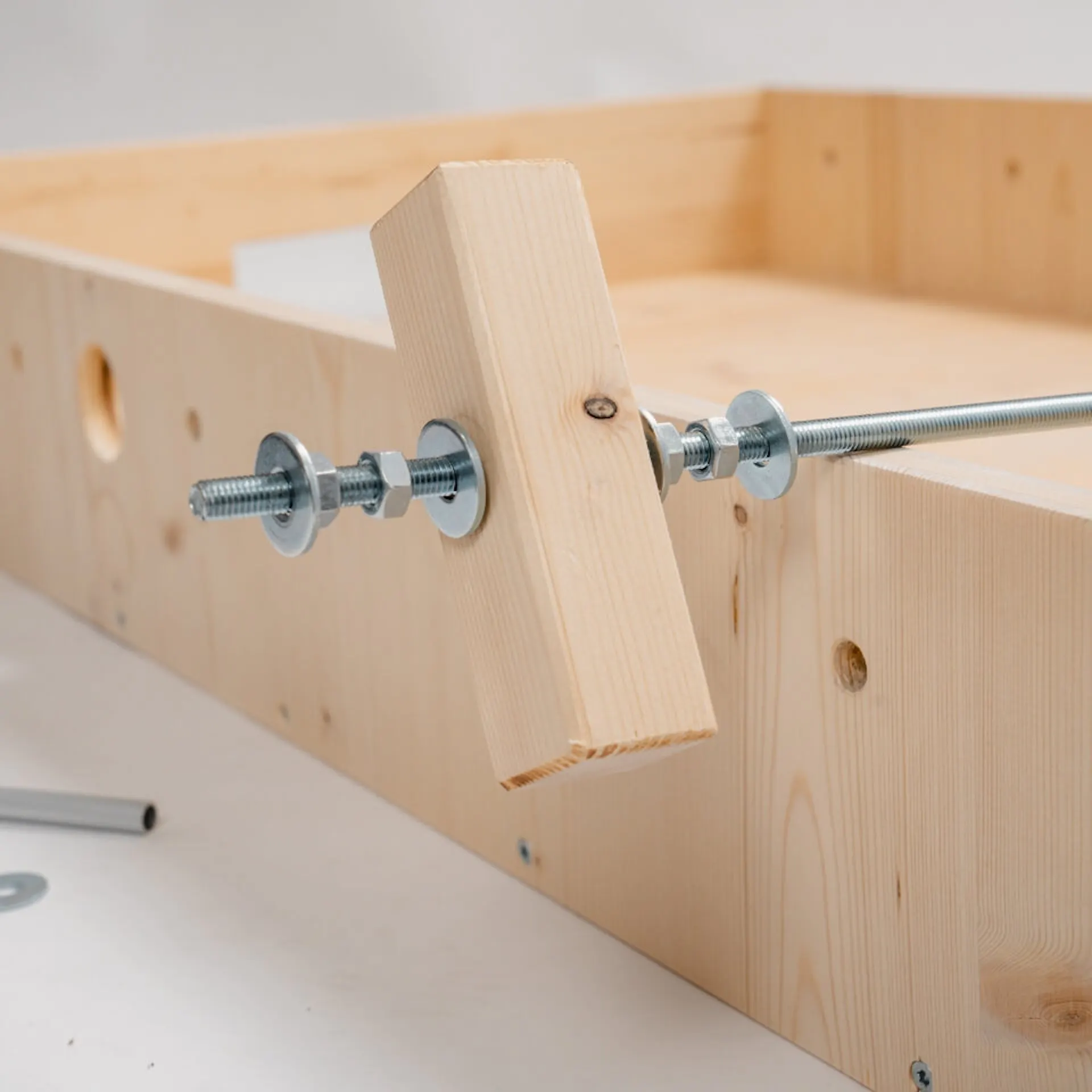 Závitová tyč, na ní hranaté dřevo a několik matic a podložek. Toto je na dřevěné konstrukci. Před ním je hliníková trubka, matice a podložka.