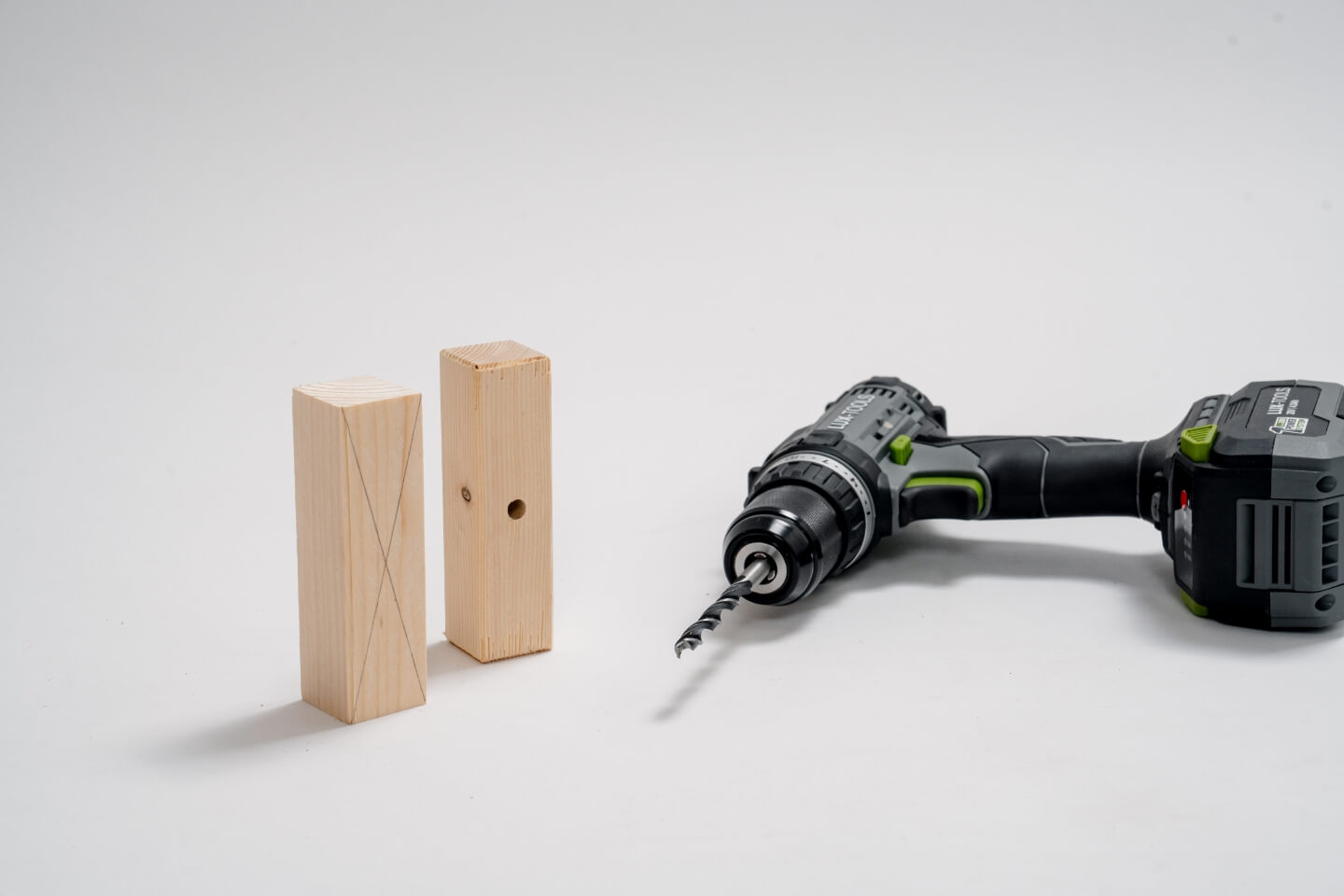 Dva hraněné trámy, na jednom nakreslený kříž, na druhém nastojato vyvrtaný otvor. Vedle je akumulátorový šroubovák.