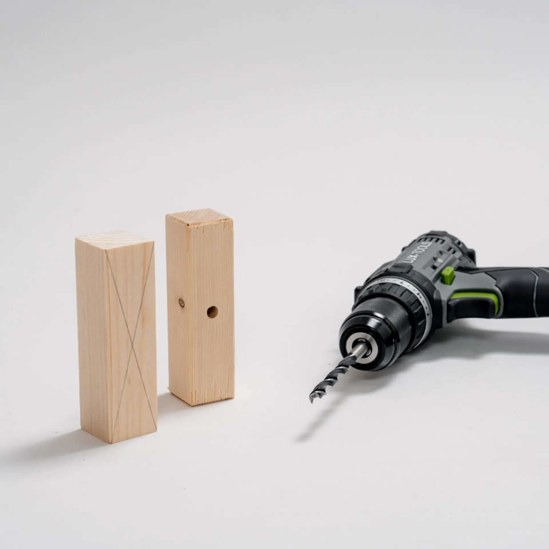 Dva hraněné trámy, na jednom nakreslený kříž, na druhém nastojato vyvrtaný otvor. Vedle je akumulátorový šroubovák.