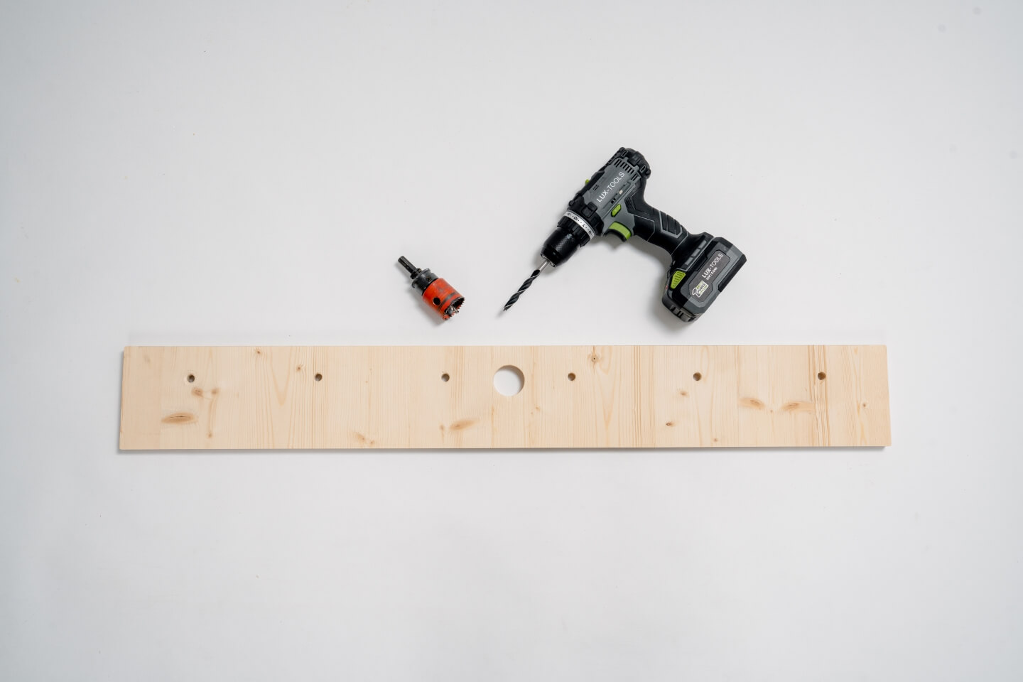 dřevěná deska s větším otvorem uprostřed, vedle jsou na každé straně tři malé otvory. Za deskou je akumulátorový šroubovák a vrtačka.