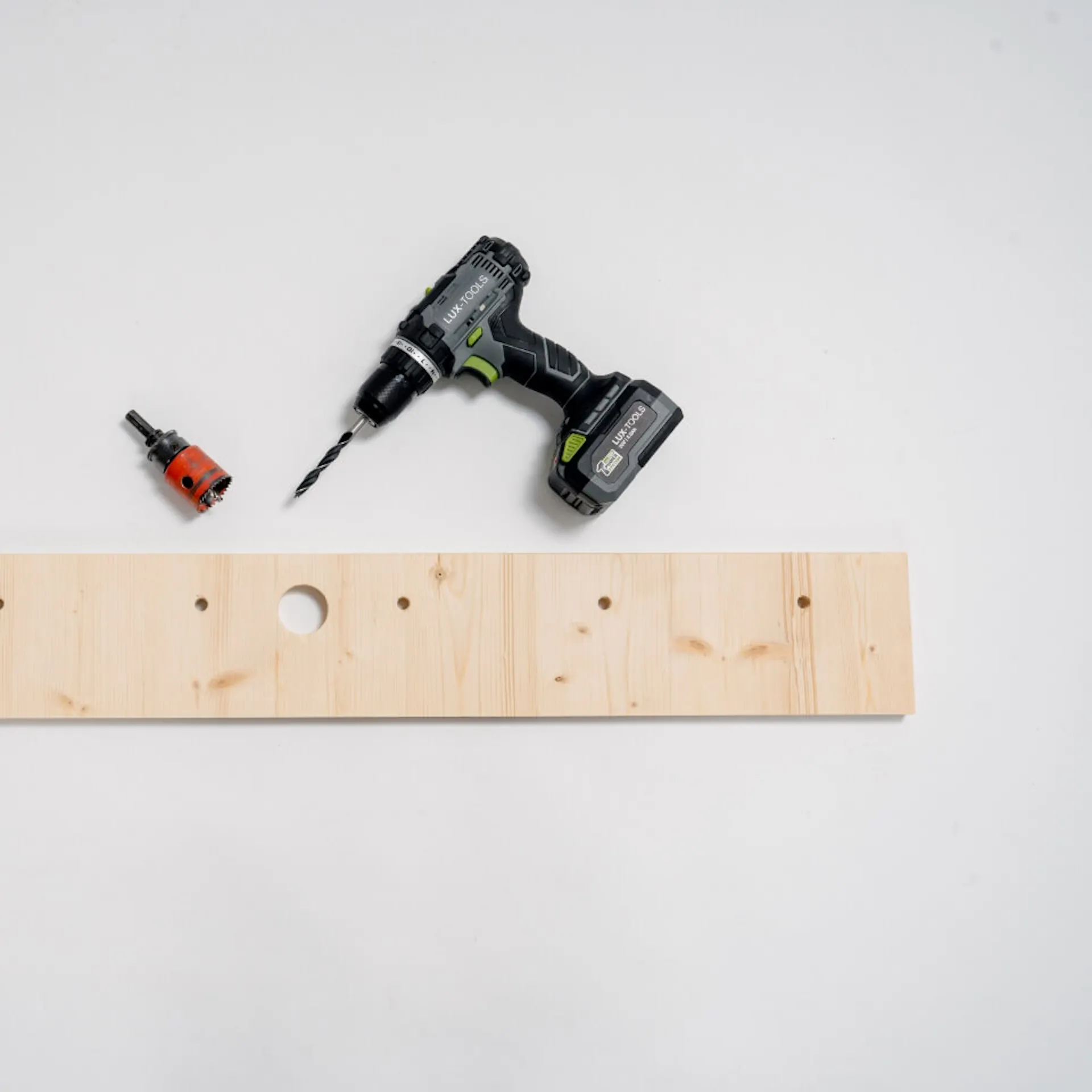 dřevěná deska s větším otvorem uprostřed, vedle jsou na každé straně tři malé otvory. Za deskou je akumulátorový šroubovák a vrtačka.