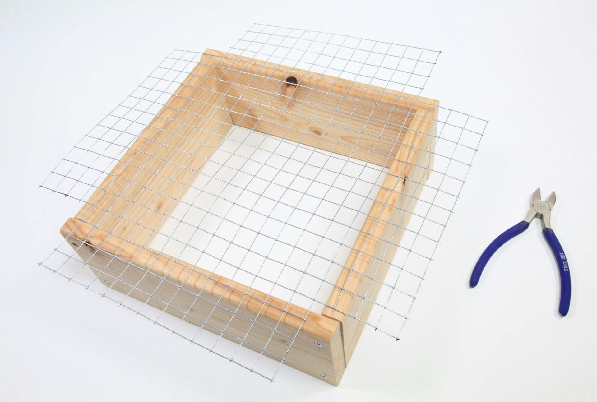 Itt egy fakeret látható, amelyen kereszt alakban drótháló van kifeszítve. Mellette egy oldalcsípőfogó található.