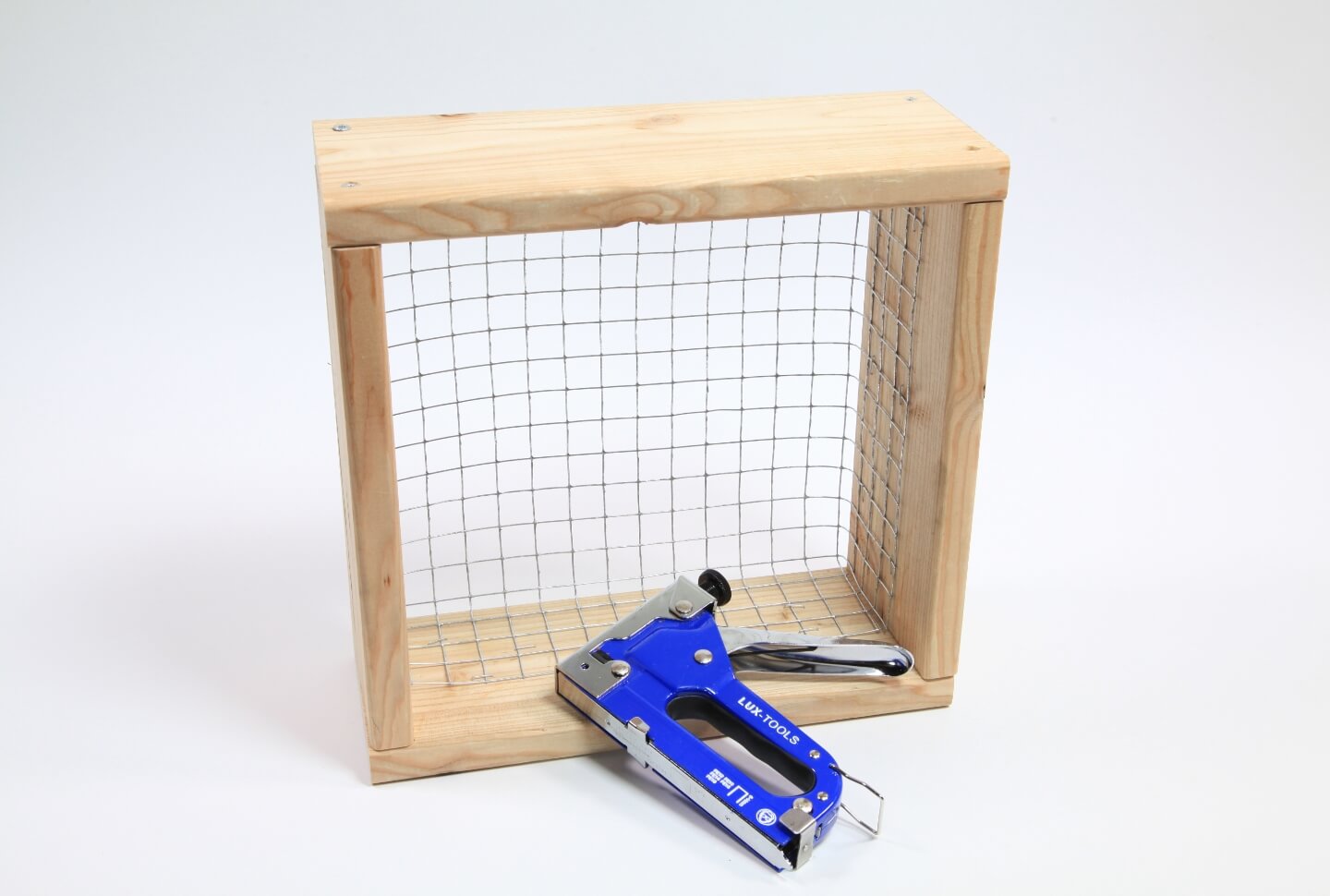 Fehér alapon a vertikális keret alapeleme, egy dróttal feszített fakeret látható. Előtte egy tűzőgép található.