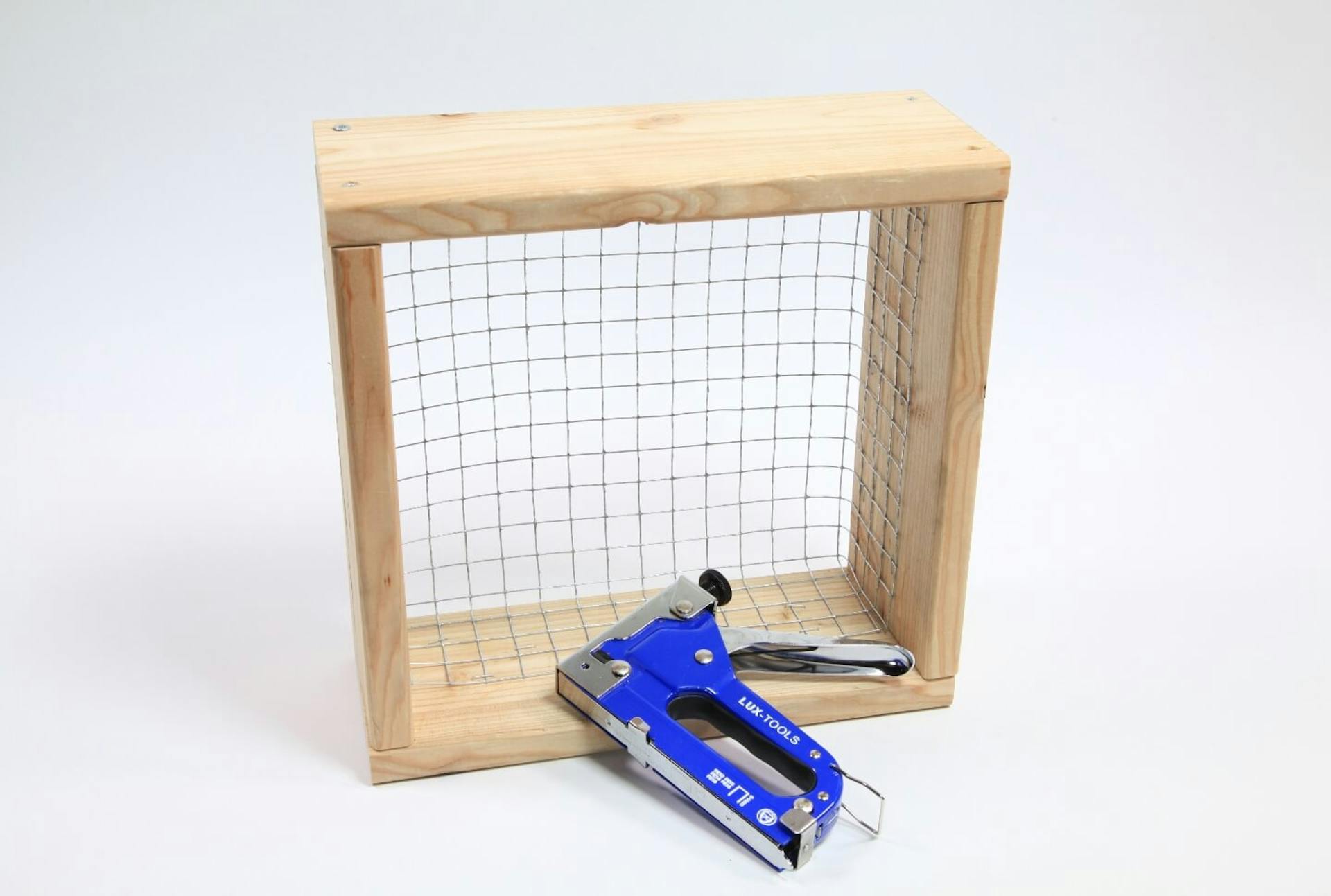 Fehér alapon a vertikális keret alapeleme, egy dróttal feszített fakeret látható. Előtte egy tűzőgép található.