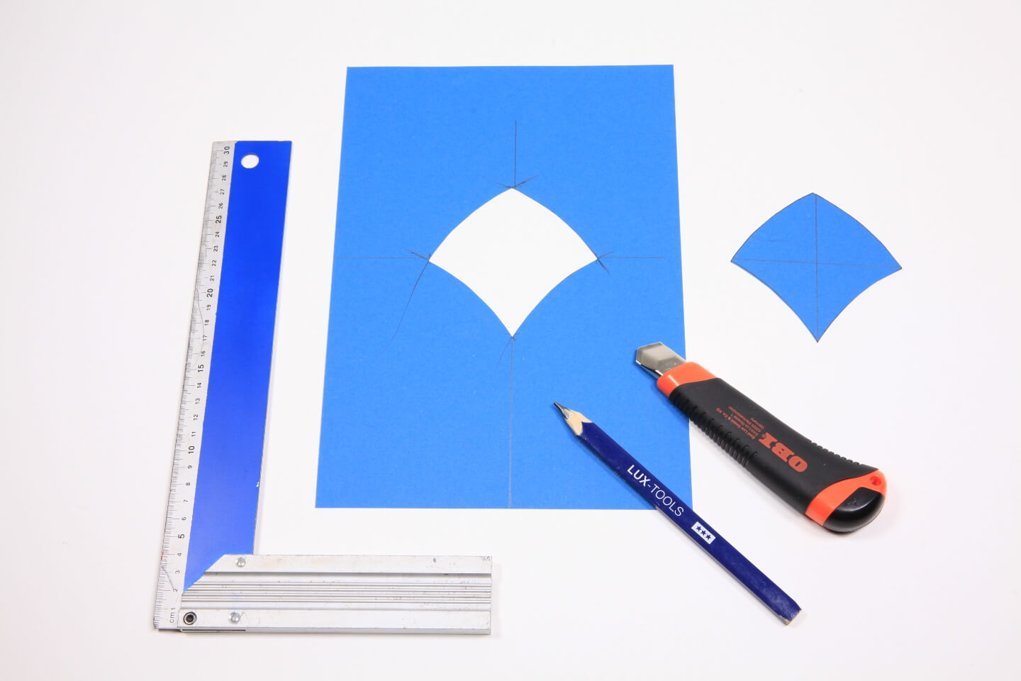 Es ist ein blaues Stück Papier zu sehen, aus welchem eine Schuppenform ausgeschnitten ist. Die Form liegt neben dem Papier. Neben dem Papier liegt zudem ein Winkelmesser. Auf dem Papier liegen ein Bleistift und ein Cutter-Messer.