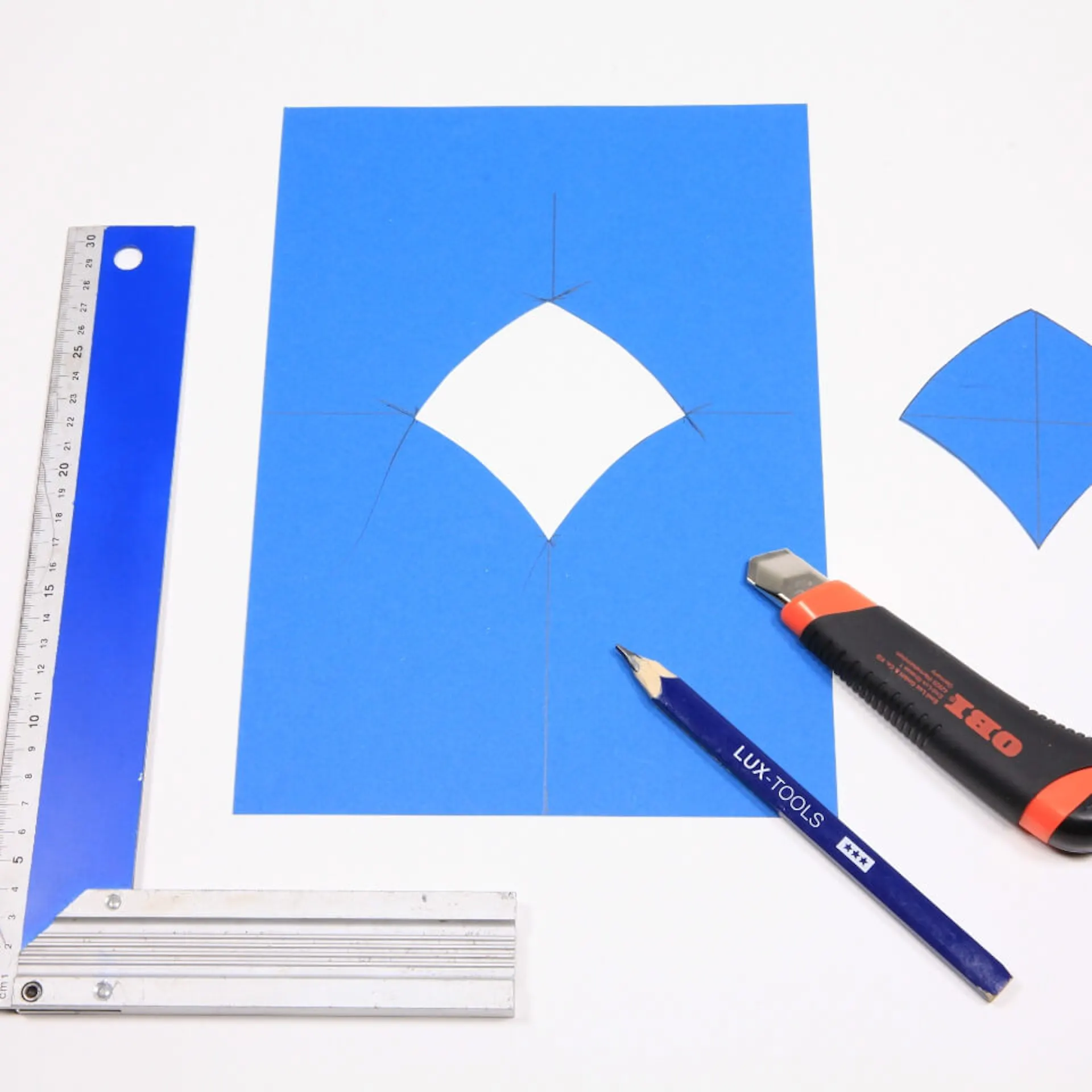 Es ist ein blaues Stück Papier zu sehen, aus welchem eine Schuppenform ausgeschnitten ist. Die Form liegt neben dem Papier. Neben dem Papier liegt zudem ein Winkelmesser. Auf dem Papier liegen ein Bleistift und ein Cutter-Messer.