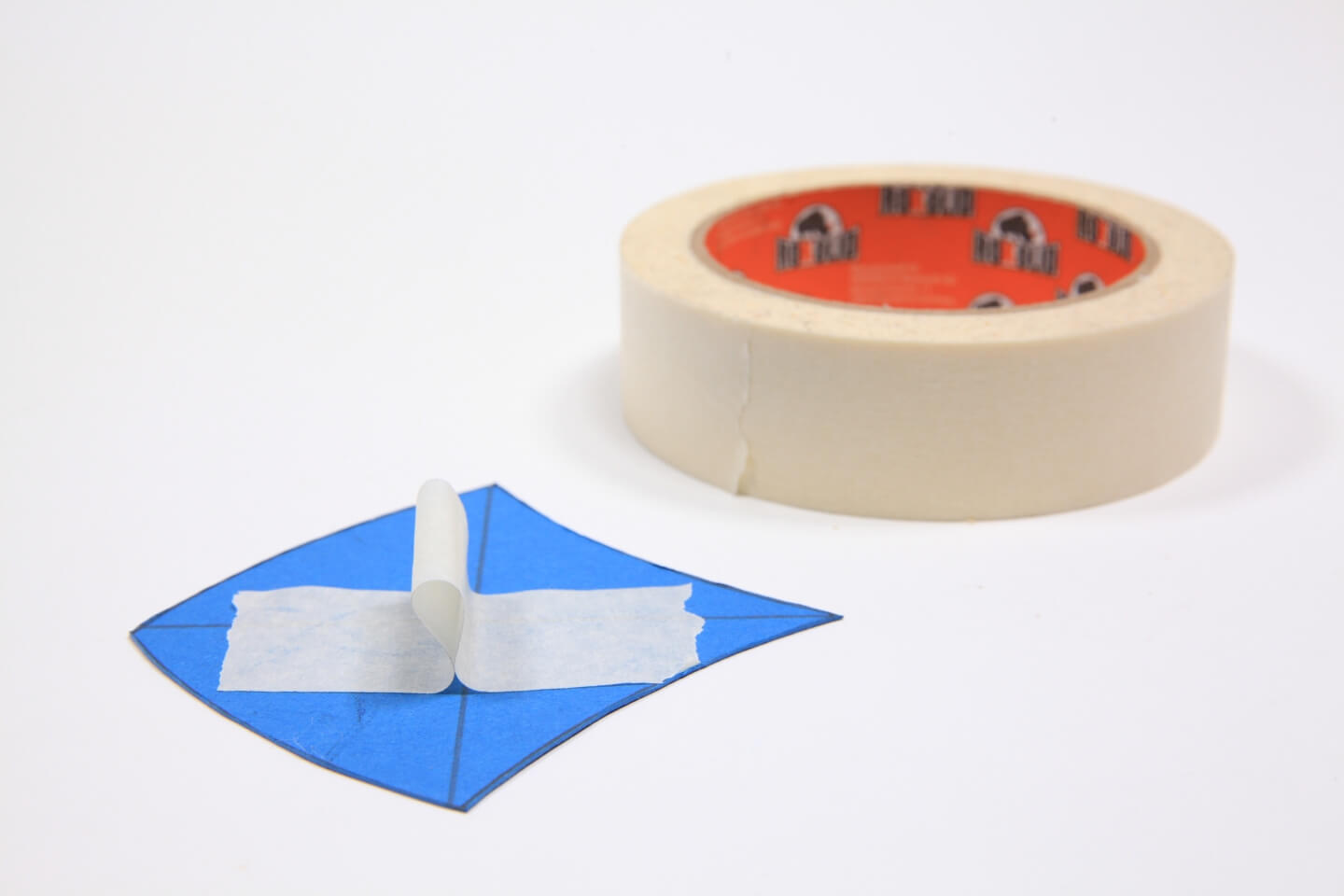Es ist ein blaues Stück Papier in Schuppenform zu sehen. Auf der Form ist ein Stück Kreppband befestigt. Im Hintergrund liegt eine Rolle Kreppband.