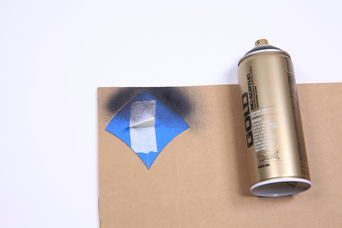 Es ist ein Stück Pappe zu sehen. Auf der oberen Ecke der Pappe liegt ein blaues Stück Papier in Schuppenform. Auf der Pappe und der Papierform ist etwas dunkle Farbe zu sehen. Daneben liegt eine goldene Sprühdose.