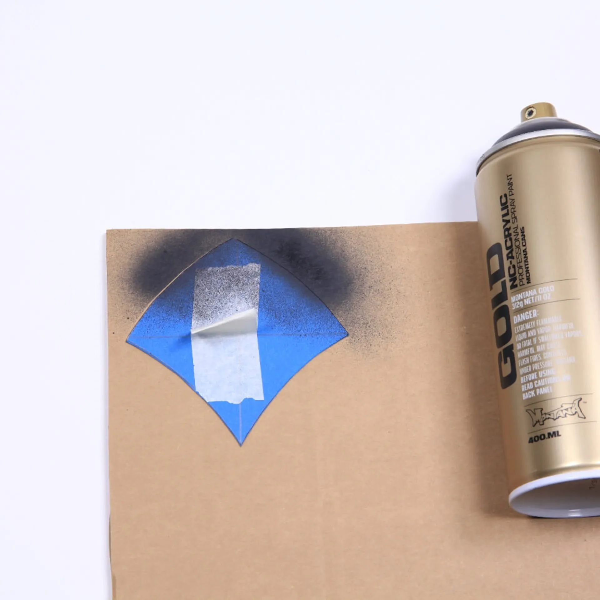 Es ist ein Stück Pappe zu sehen. Auf der oberen Ecke der Pappe liegt ein blaues Stück Papier in Schuppenform. Auf der Pappe und der Papierform ist etwas dunkle Farbe zu sehen. Daneben liegt eine goldene Sprühdose.