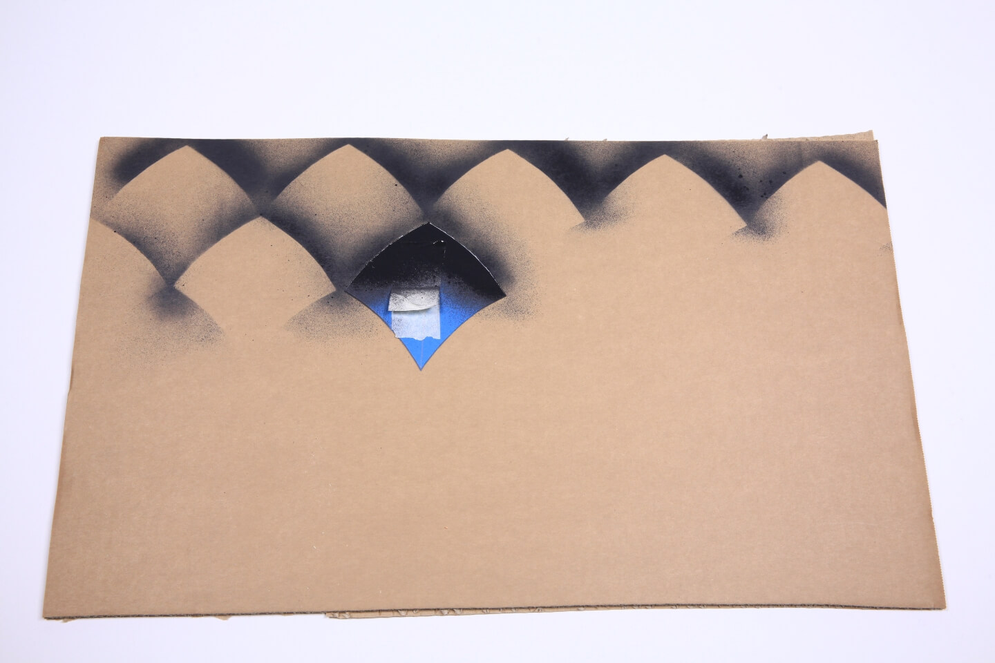 Es ist ein Stück Pappe zu sehen, welches fast bis zur Hälfte mit einem dunklen Schuppenmuster bedeckt ist. Auf der Pappe liegt ein blaues Stück Papier in Schuppenform.