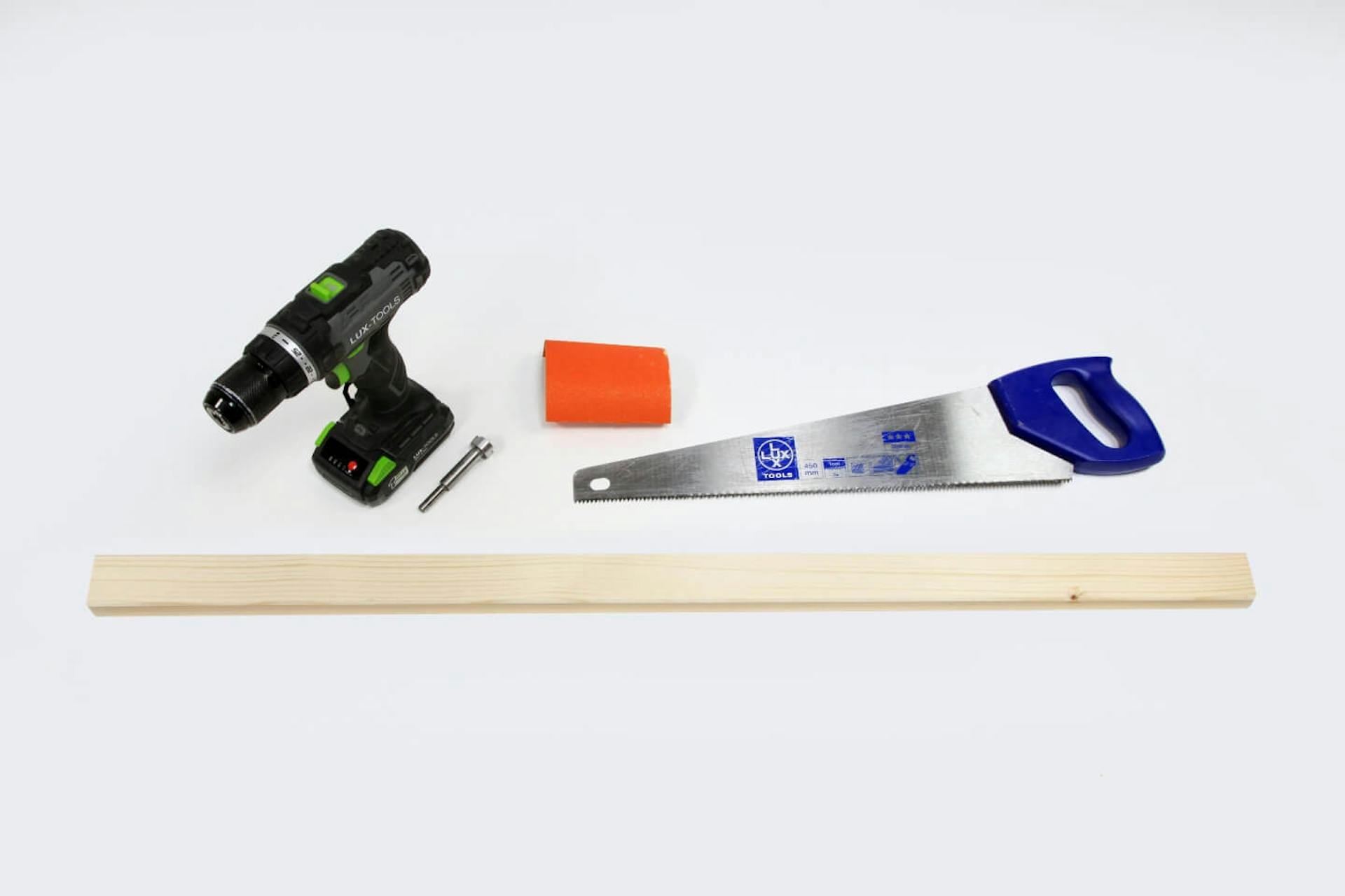 Una trave in legno su fondo bianco. Sopra la trave si trova un avvitatore a batteria. Di fianco a questo, una sega a mano e un pezzo di carta abrasiva.