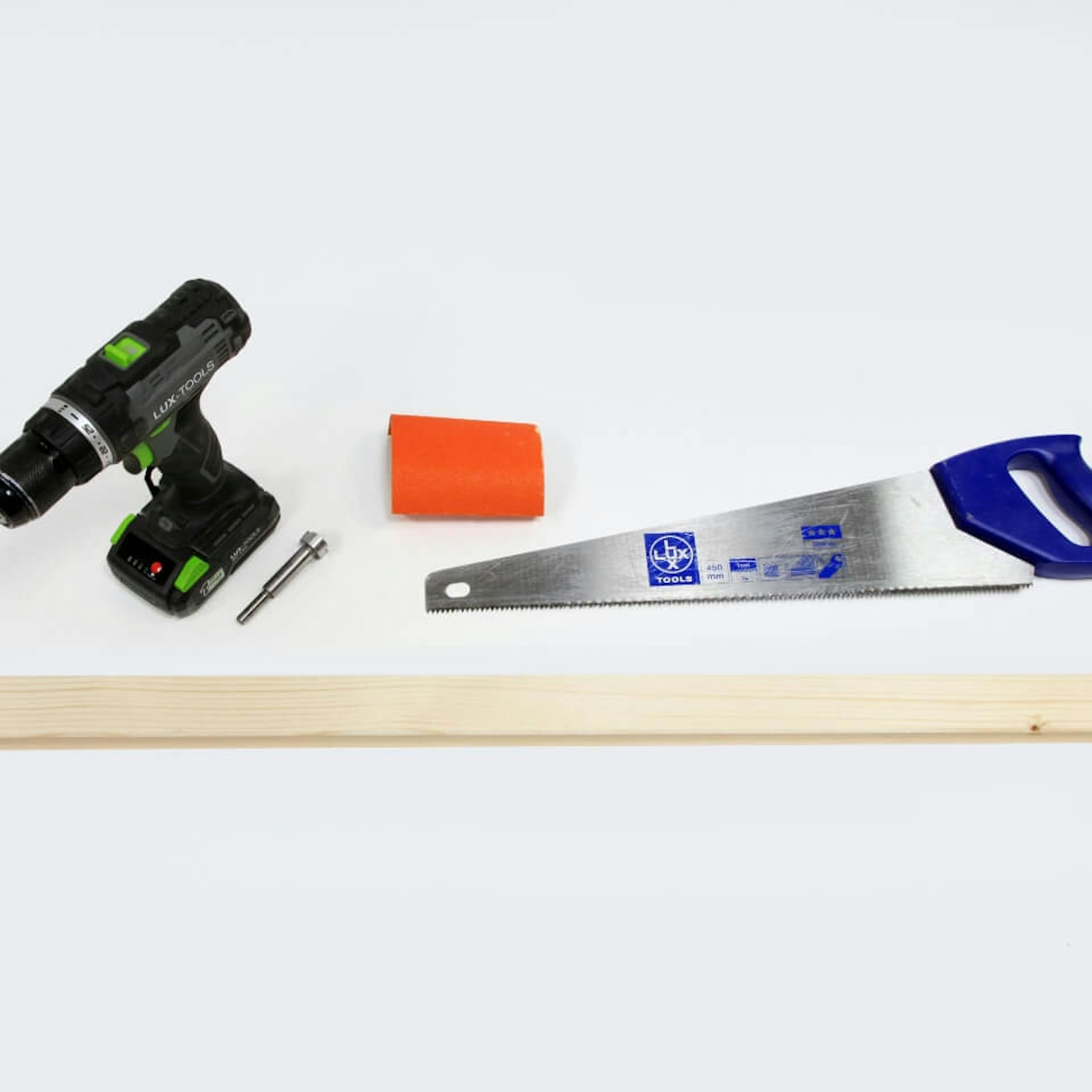 Auf einem weißen Untergrund liegt ein Kantholz. Oberhalb des Kantholzes steht ein Akkuschrauber. Daneben liegen eine Handsäge sowie ein Stück Schleifpapier.