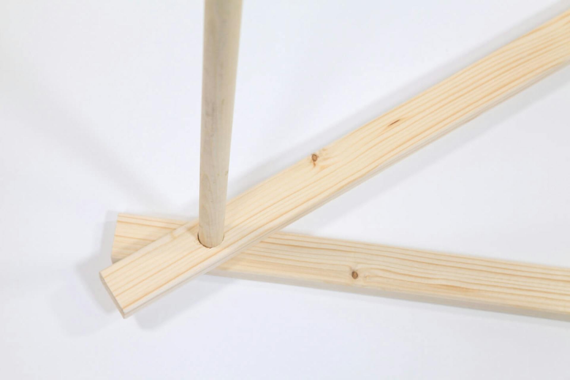 Su sfondo bianco, due travi sovrapposte alle estremità. I fori delle travi sono allineati e in essi è inserita una barra di legno che sporge verso l’alto.