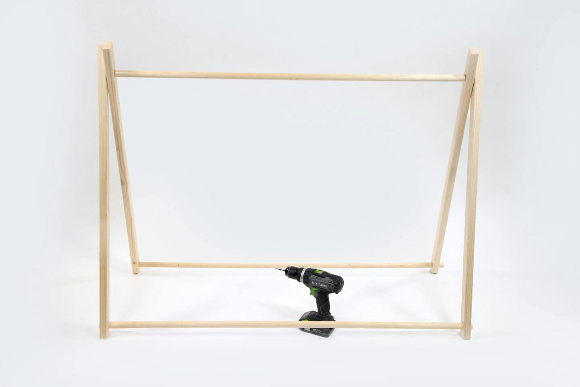 Telaio in legno di una tenda su sfondo bianco. Al centro del telaio si trova un avvitatore a batteria.