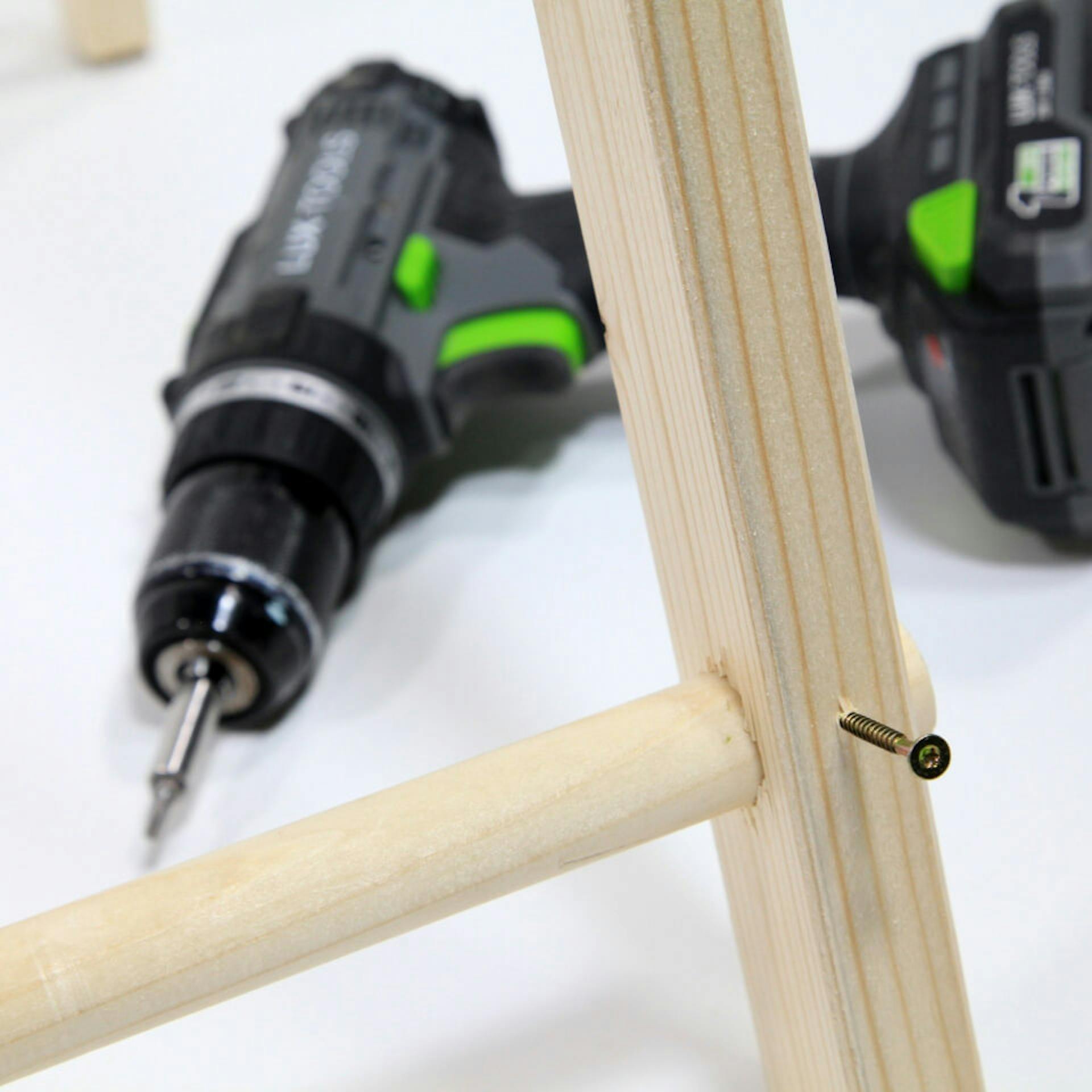 Es ist der Fuß eines Zeltgerüstes aus Holz zu sehen. Zwei Holzelemente sind durch eine Schraube verbunden. Im Hintergrund ist ein Akkuschrauber zu sehen.