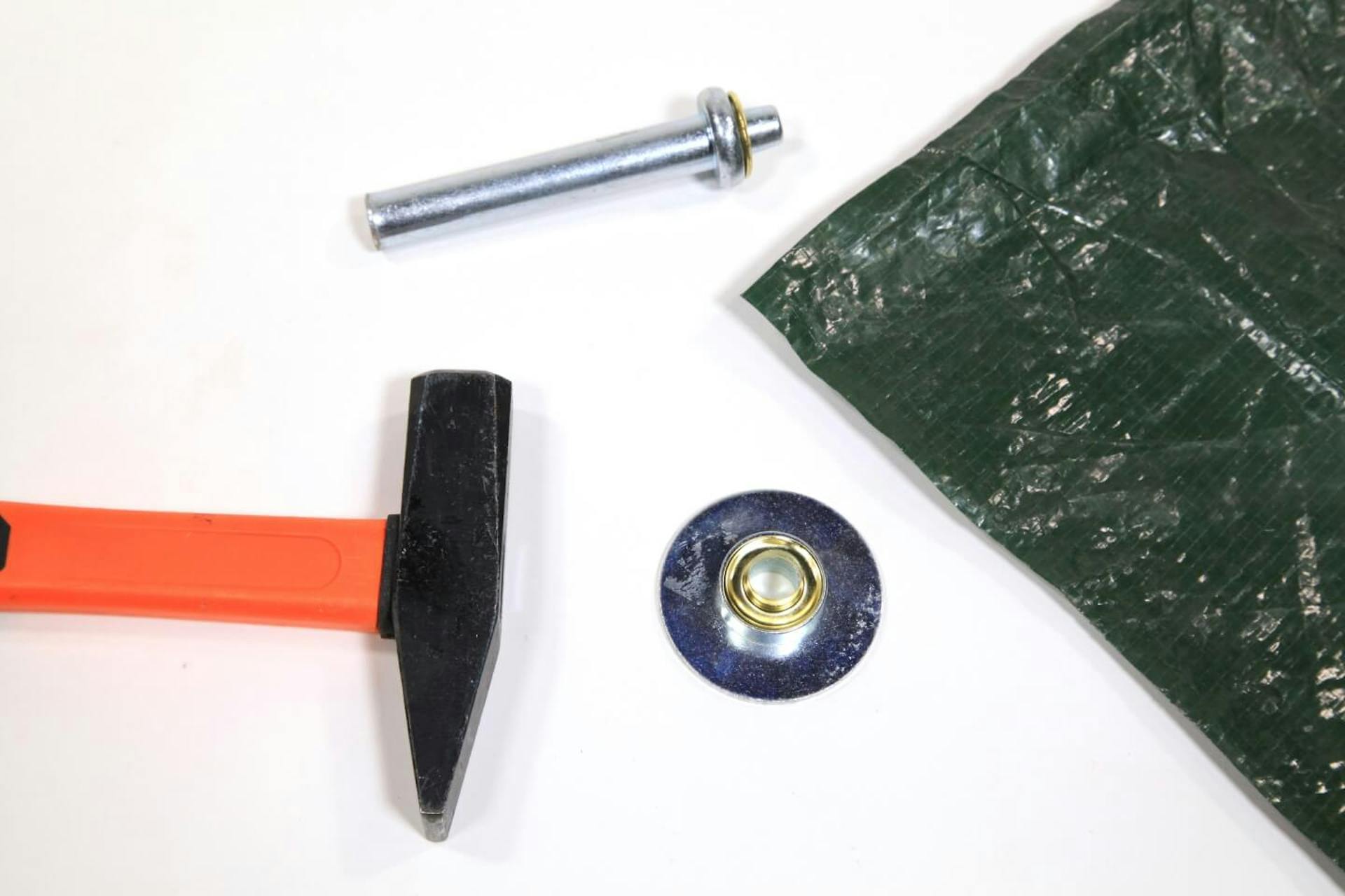 Su sfondo bianco è visibile l’angolo di un telo. Accanto a questo, un utensile per l'applicazione degli occhielli e un martello.