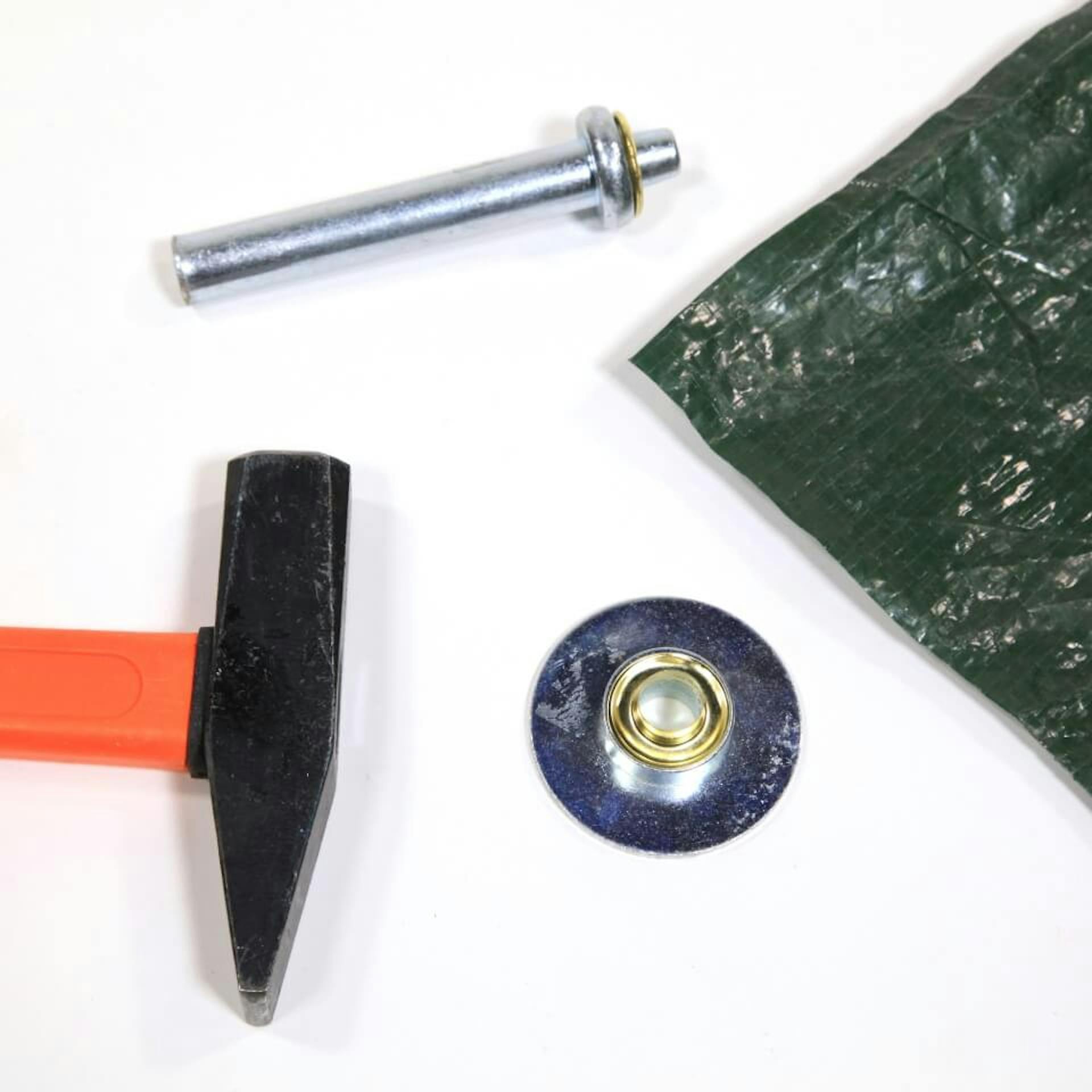 Auf einem weißen Untergrund ist die Ecke einer dunklen Plane zu sehen. Daneben liegen Werkzeug für Ösen sowie ein Hammer..