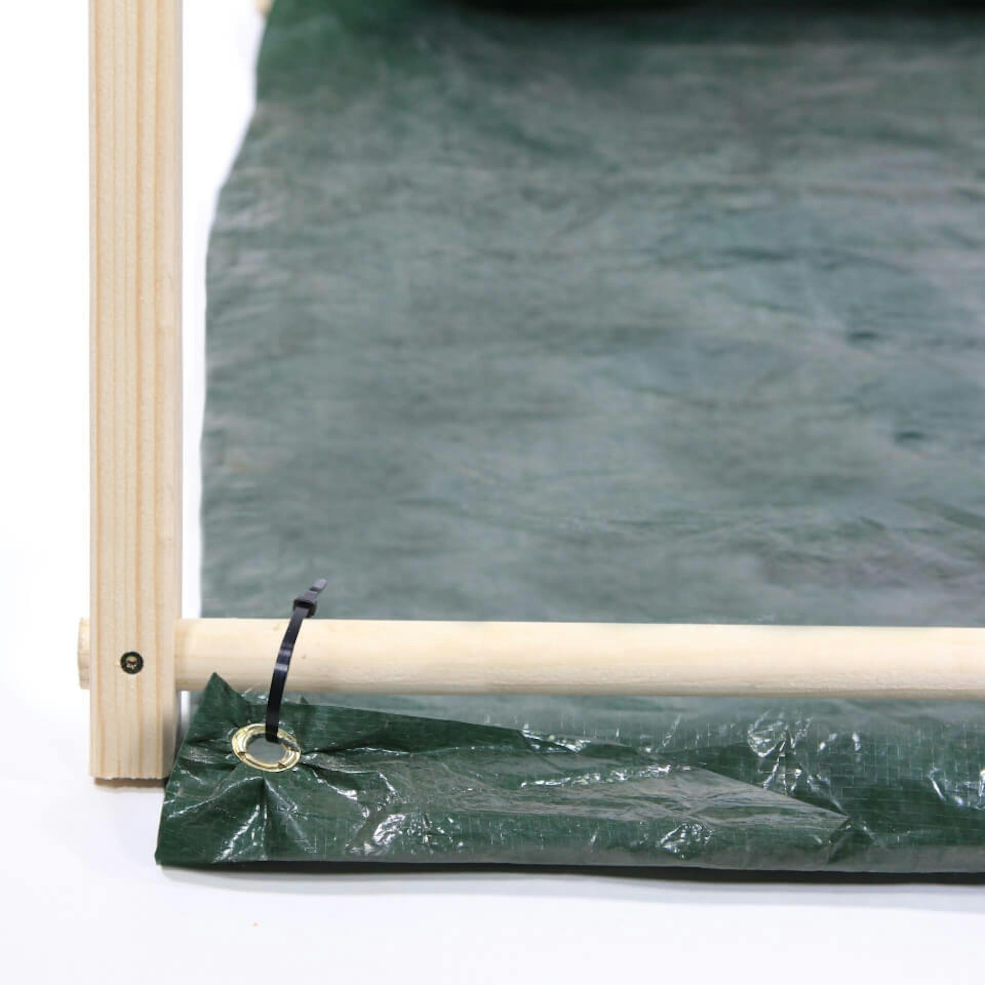 Es sind zwei miteinander verbundene Holzelemente eines Zeltgerüstes zu sehen. An einem Holzstab ist eine Plane mit Hilfe eines Kabelbinders befestigt.