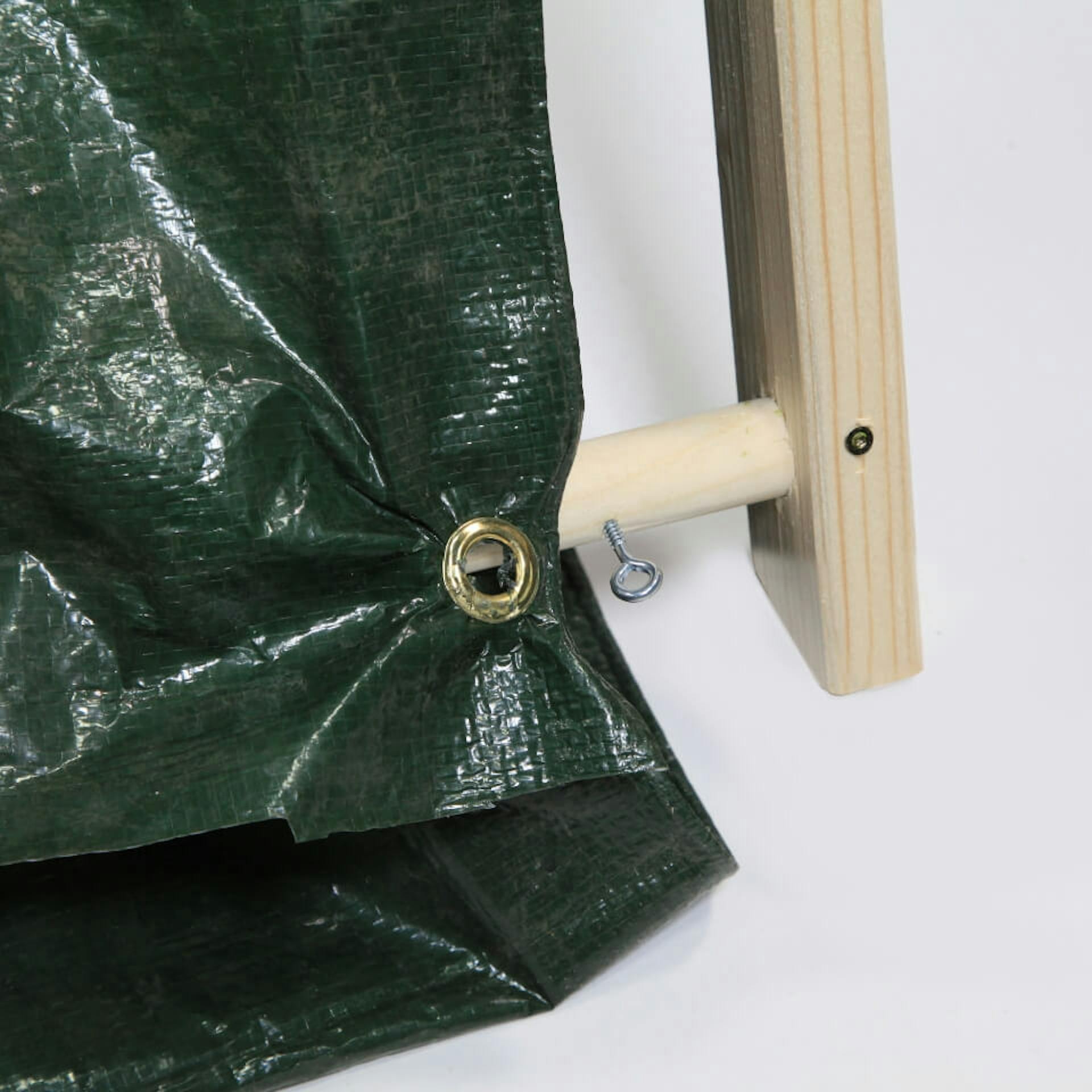 Auf einem weißen Untergrund ist der Fuß eines Zeltgerüstes aus Holz zu sehen. In einer Holzstange steckt eine Schraubenöse. An der Vorderseite des Gerüstes ist eine dunkle Plane befestigt.