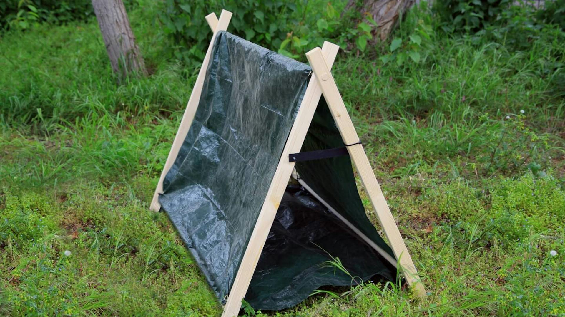 Es ist ein kleines Zelt zu sehen, welches auf einer Wiese steht. Das Gerüst besteht aus Kanthölzern, die Abdeckung aus einer dunklen Plane. Im Hintergrund sind Baumstämme zu erkennen.