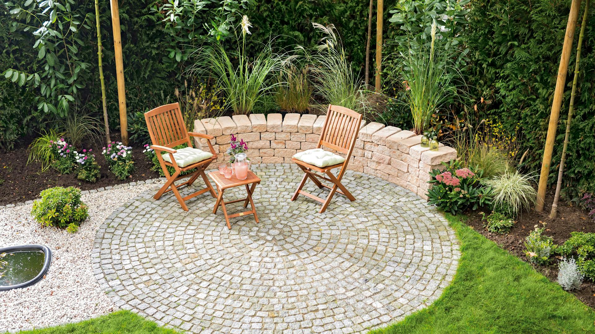 Ideen für die Gartengestaltung mediterraner Sitzplatz mit Holzmöbeln auf Pflastersteinen an Sandsteinmauer umgeben von mediterraner Bepflanzung