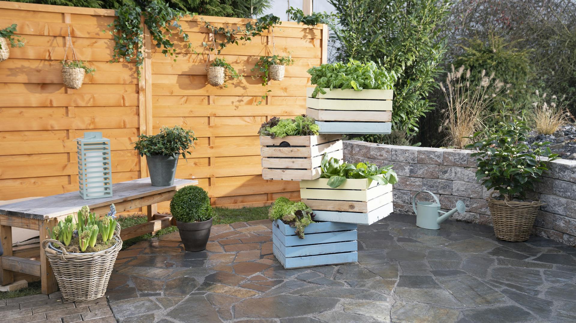 Ein fertiger Turm aus Holzkisten steht, bepflanzt mit Gemüse und Kräutern, auf einer Terrasse.