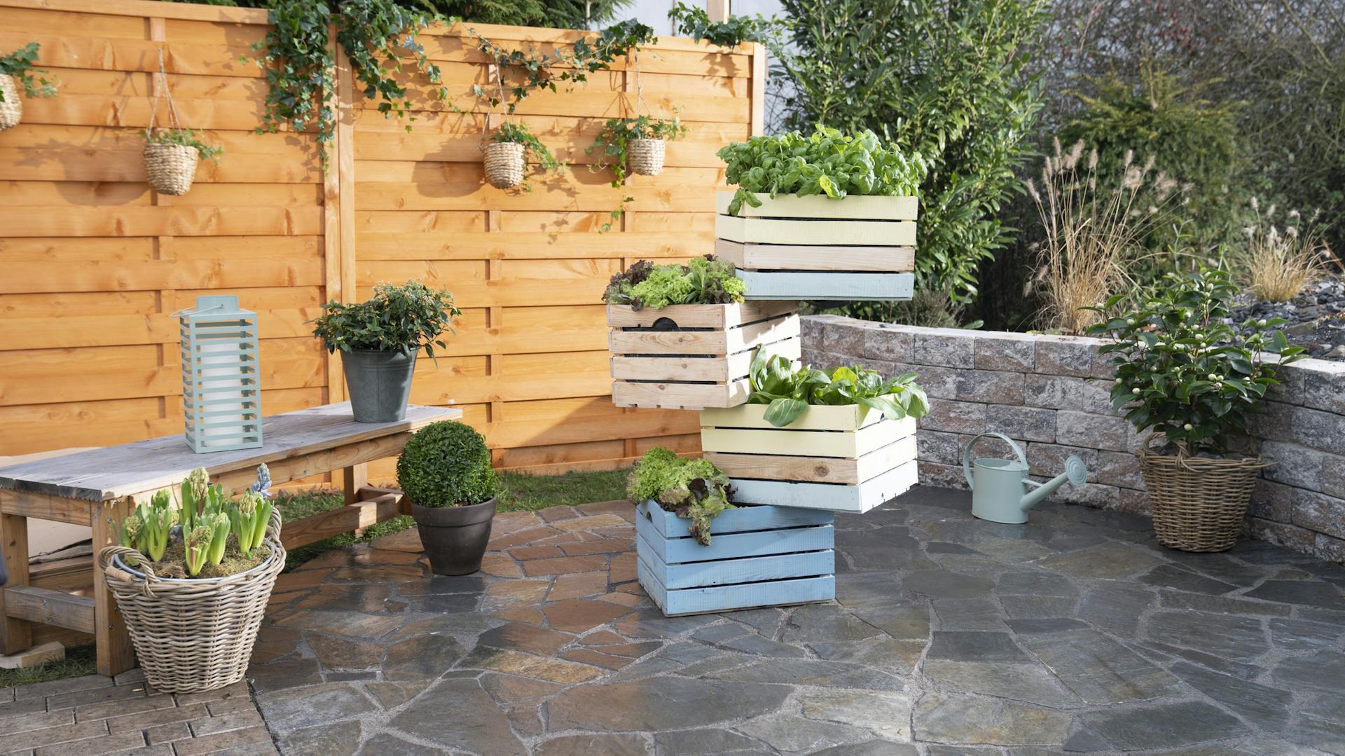 Ein fertiger Turm aus Holzkisten steht, bepflanzt mit Gemüse und Kräutern, auf einer Terrasse.