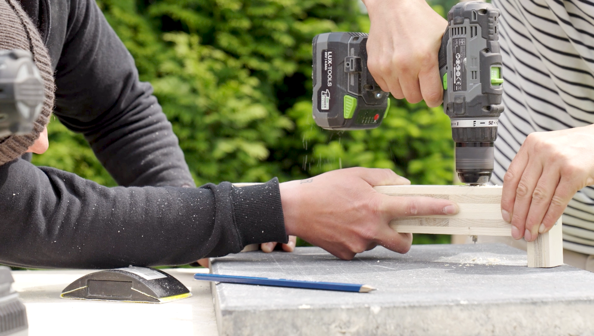 Az egyik barkácsoló férfi egy négyszöglécet tart, a másik pedig lyukat fúr a léc oldalába.