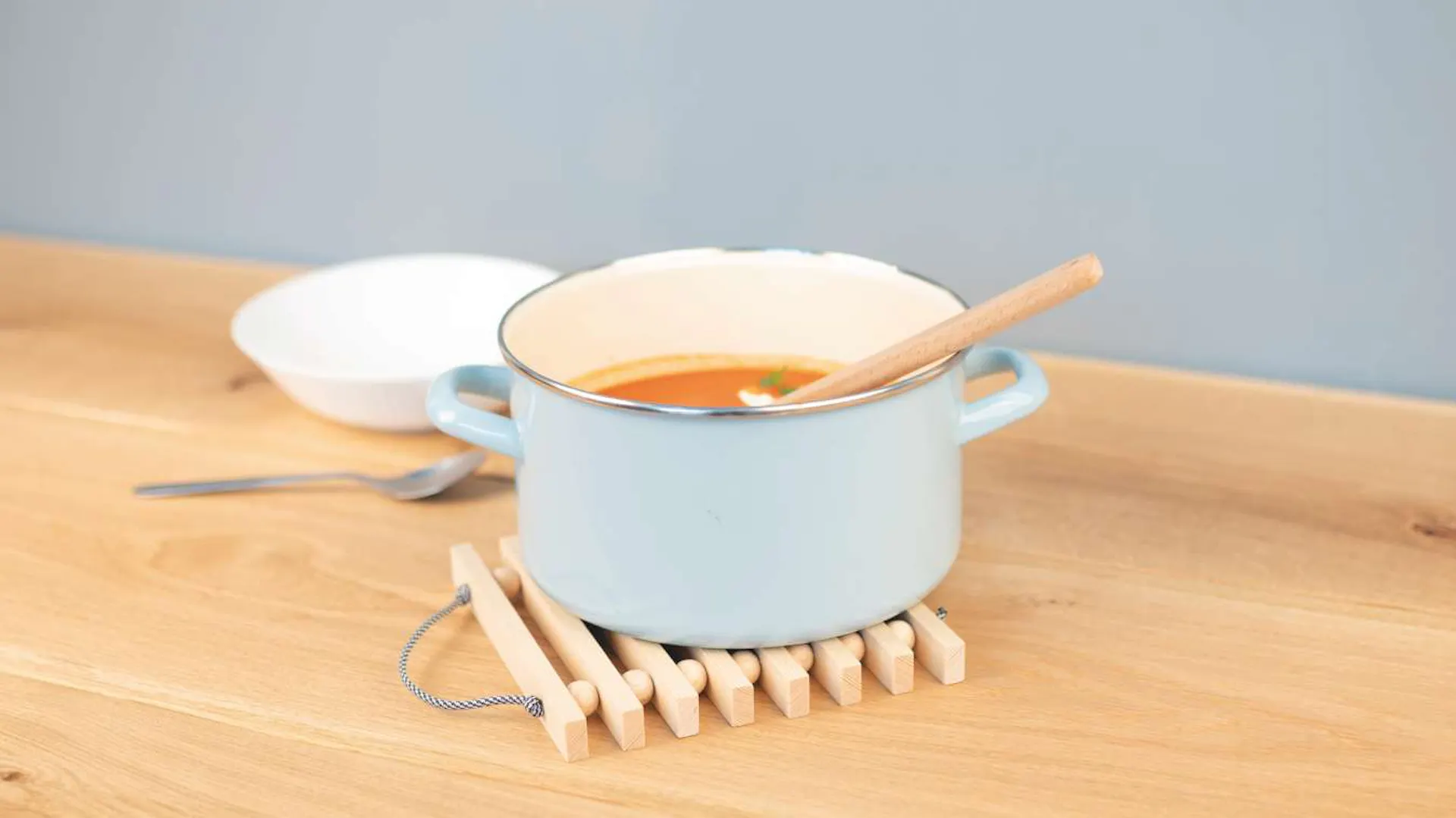 Auf einem Leistenuntersetzer steht einer kleiner, hellblauer Topf, in dem Suppe ist