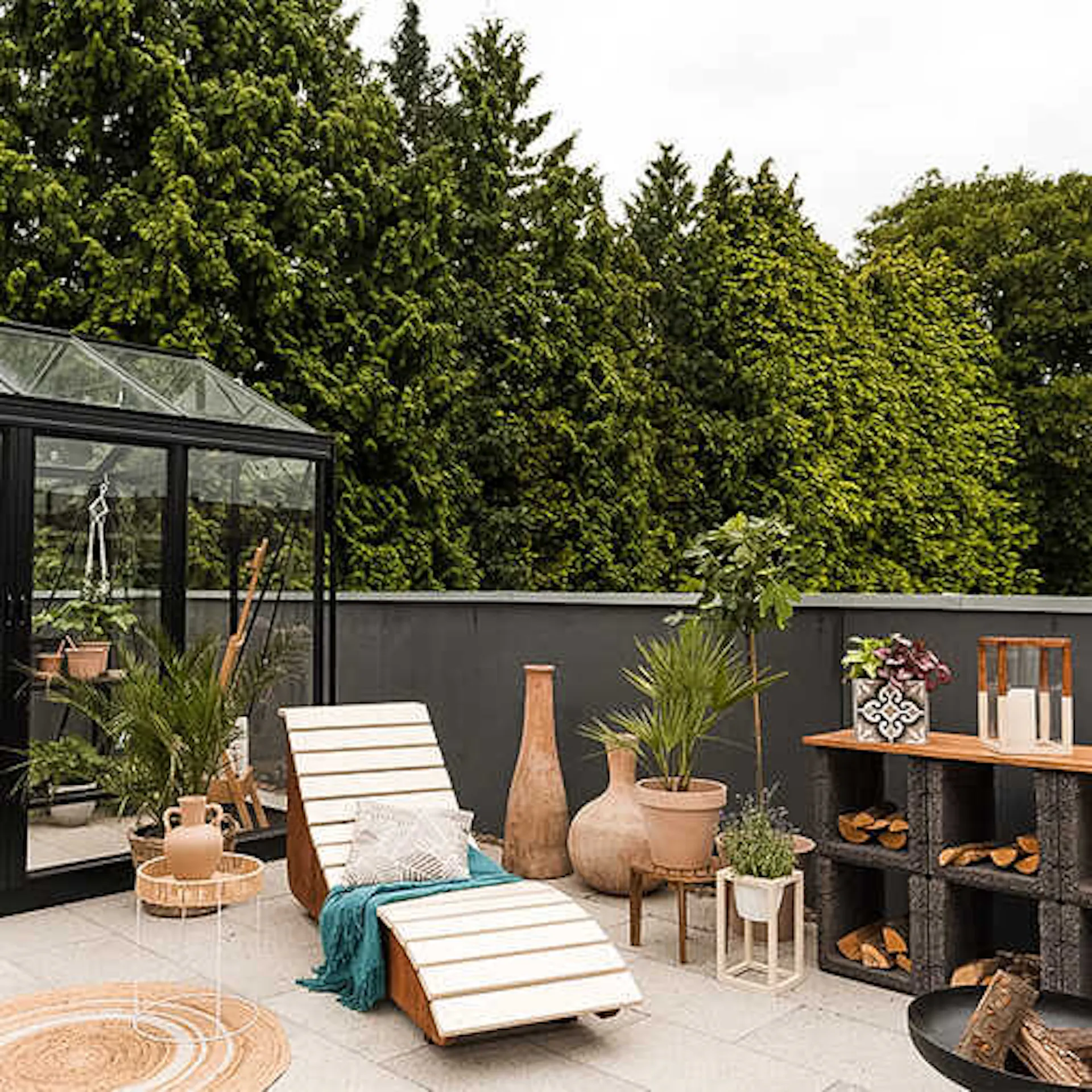 Vor einem Gewächshaus stehen auf einer Terrasse ein Beistelltisch, eine Liege und viele Pflanzen